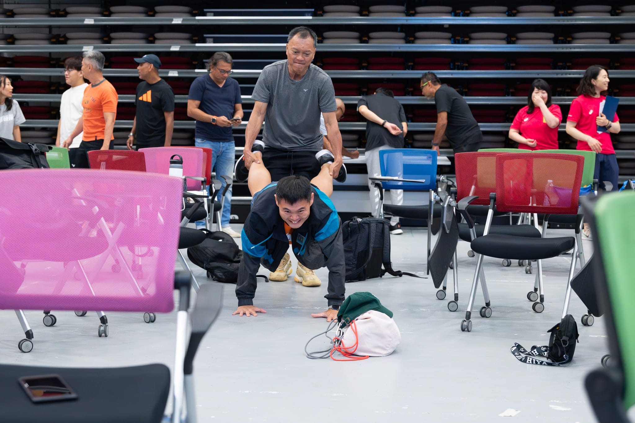 A man does a push-up position while an older man holds his legs in an indoor sports hall with colorful chairs.