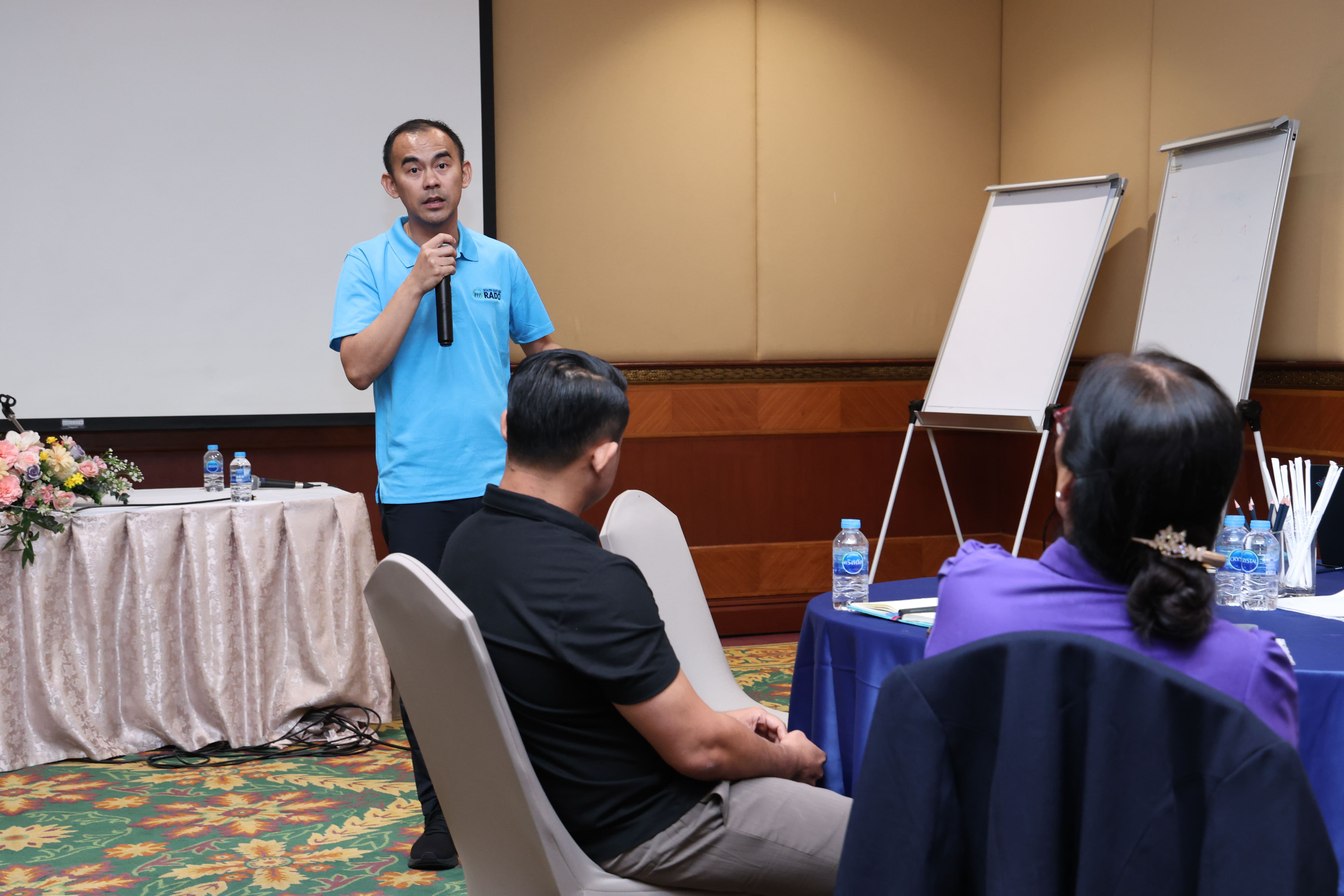 Man in blue polo holds a microphone speaking to seated attendees; flip charts and tables with water bottles nearby.