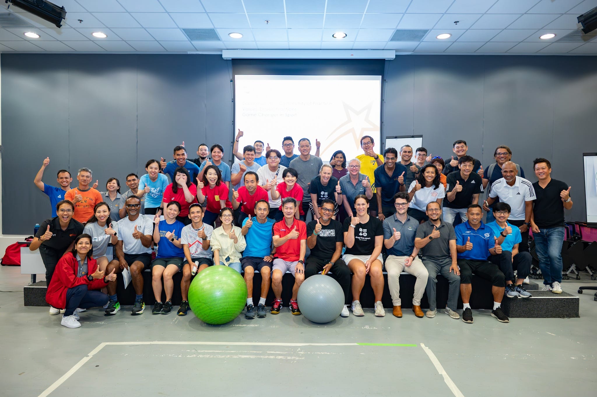 Large group of people posing with thumbs up in a room with a projection screen and two exercise balls on the floor.