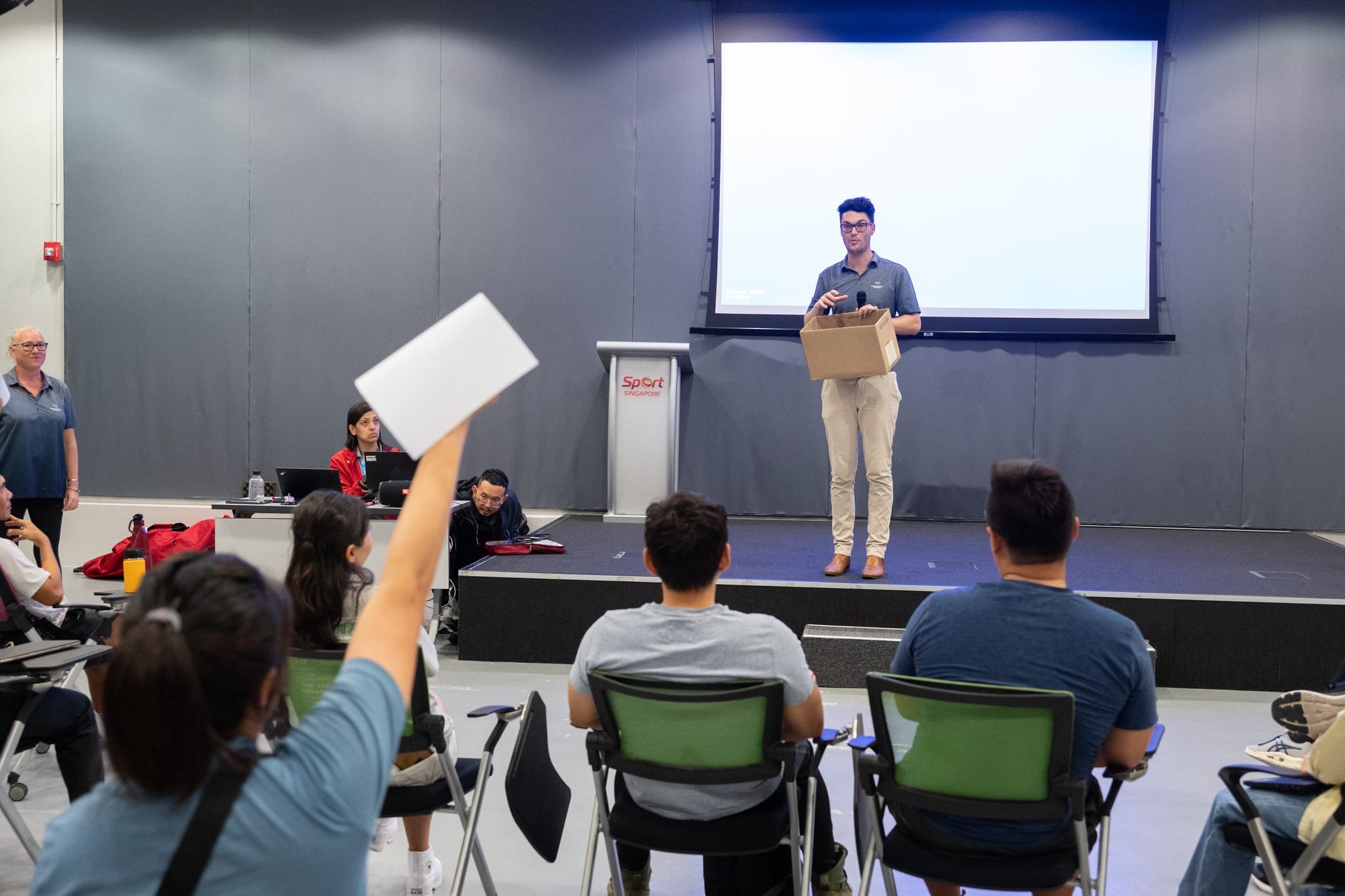 A man holds a box on stage at a Sport Singapore venue while an audience member raises a paper in the foreground.
