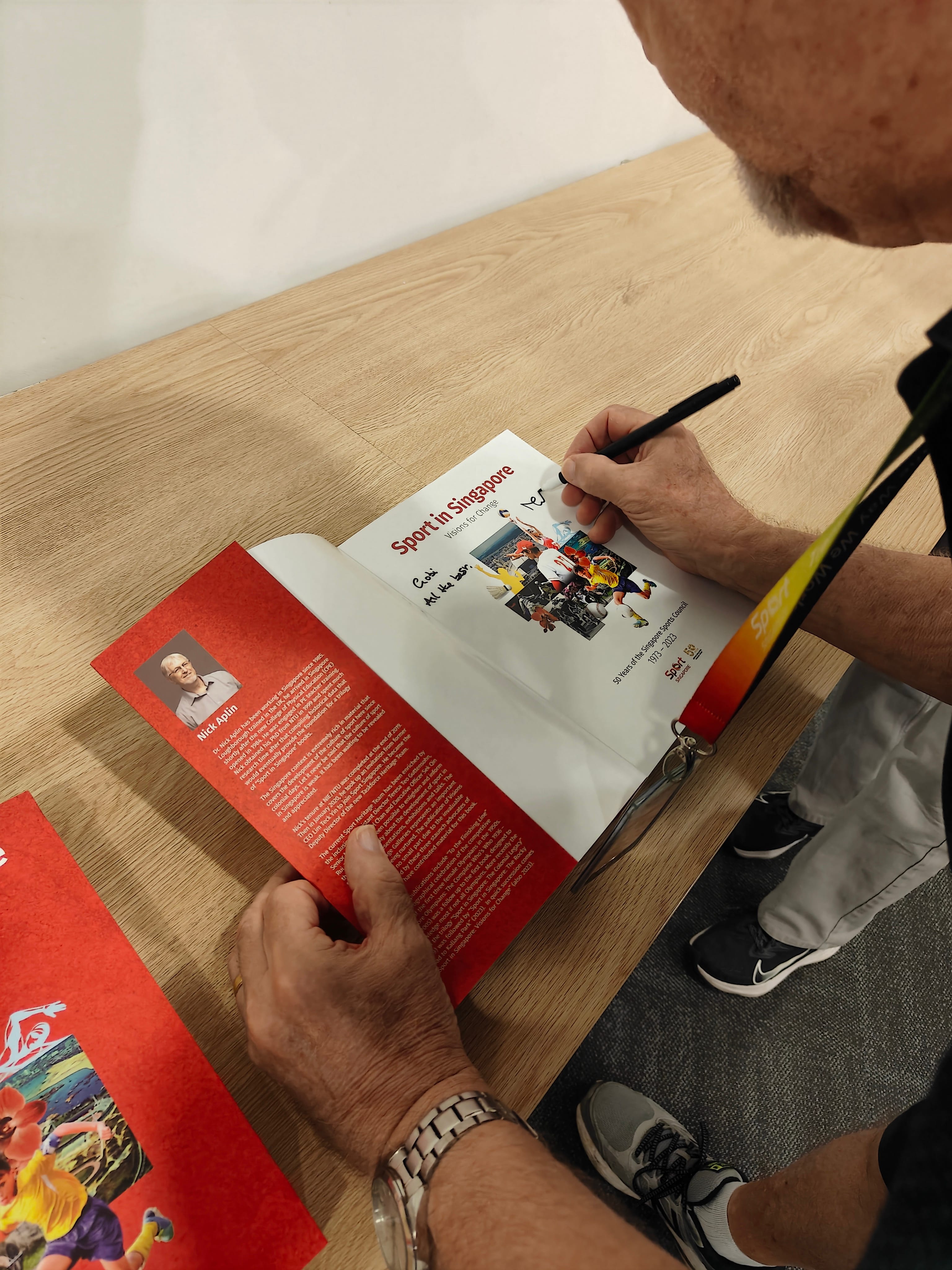 Person signs a book titled Sport in Singapore at a wooden table, holding it open to a red page.