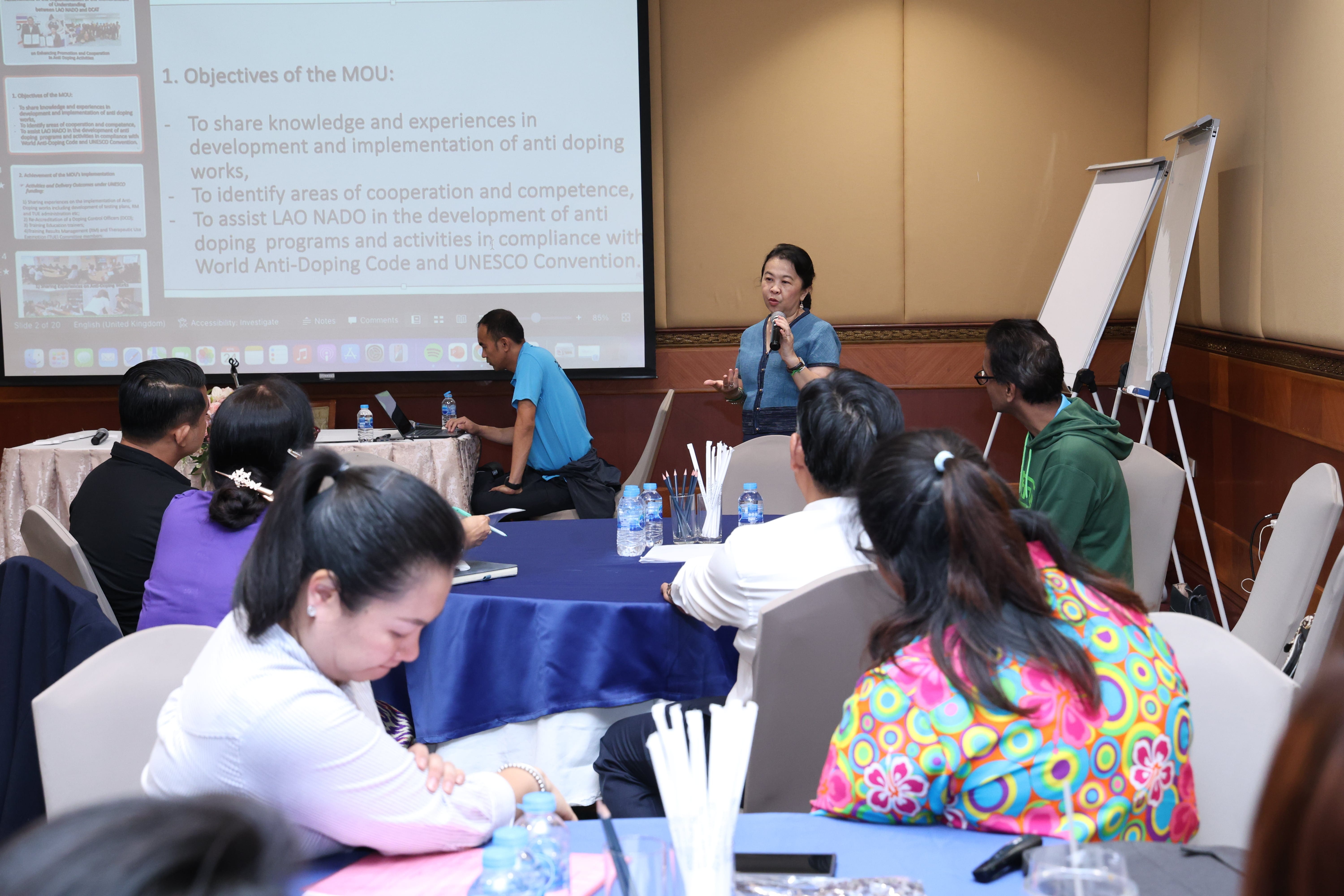 Woman speaks into a microphone to seated attendees; projector slide and flip chart in a conference room.