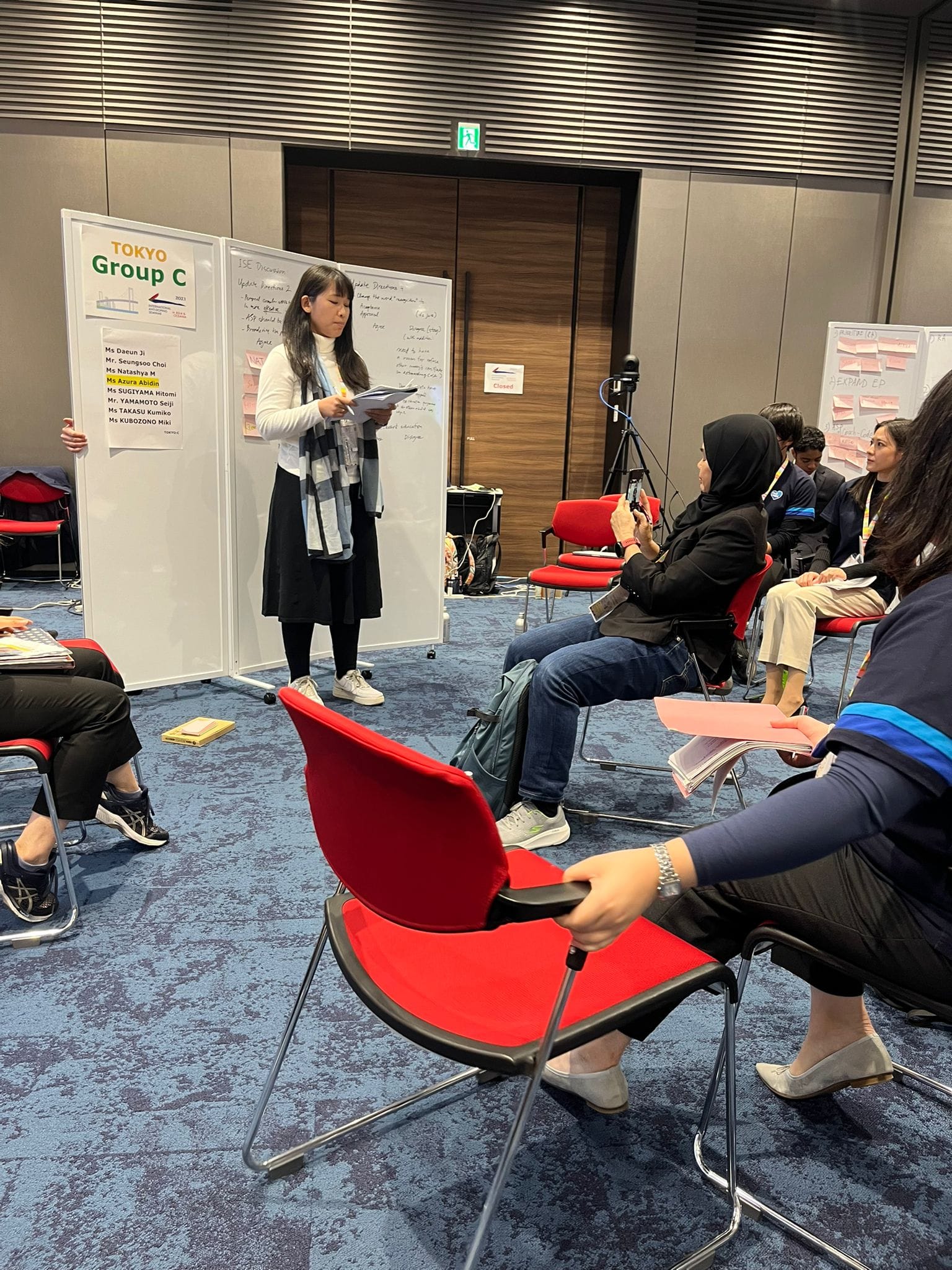 A woman presents to a seated group in a conference room beside a whiteboard labeled "Tokyo Group C."