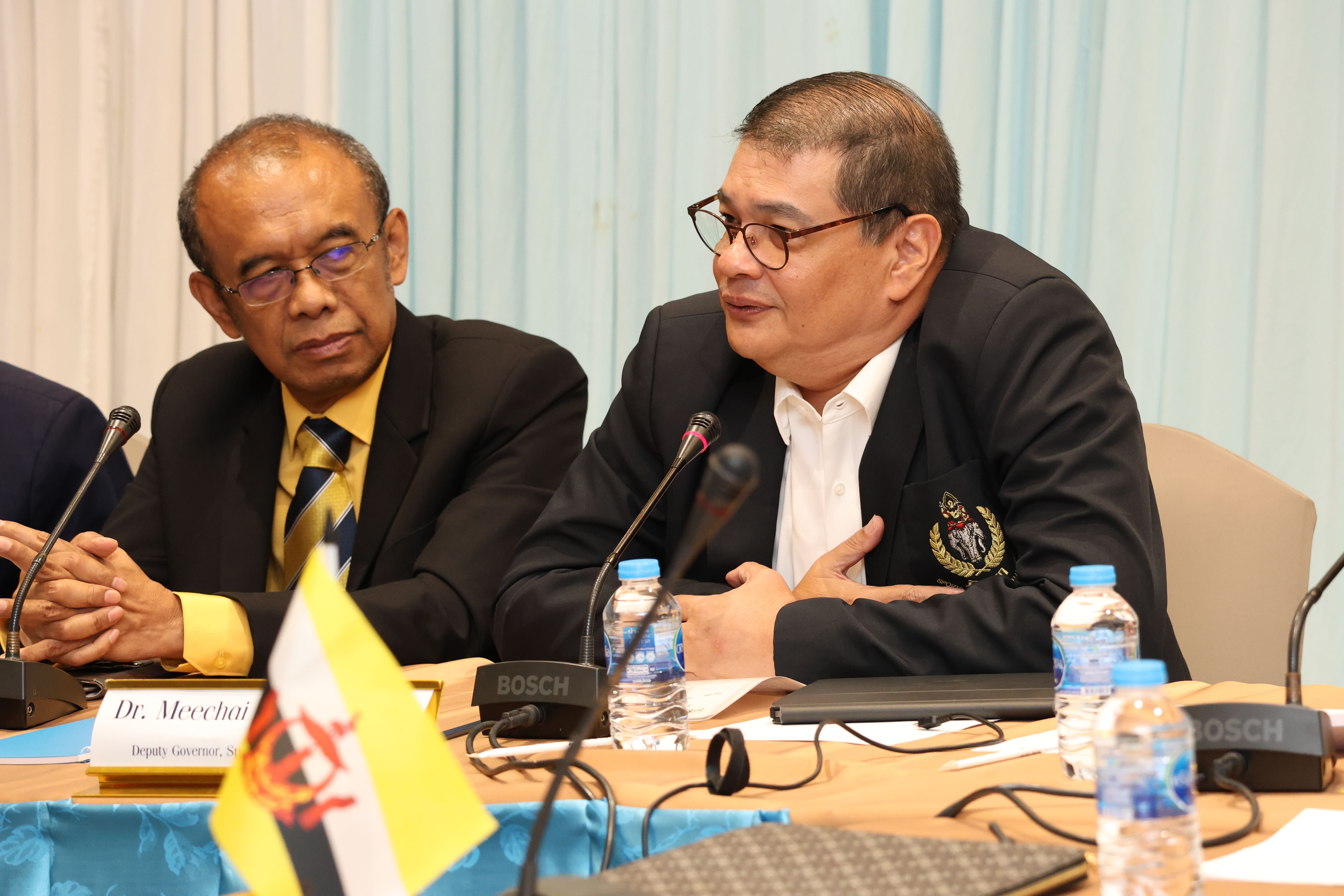 Two men in suits sit at a meeting table with microphones and water bottles; one speaks, the other listens.
