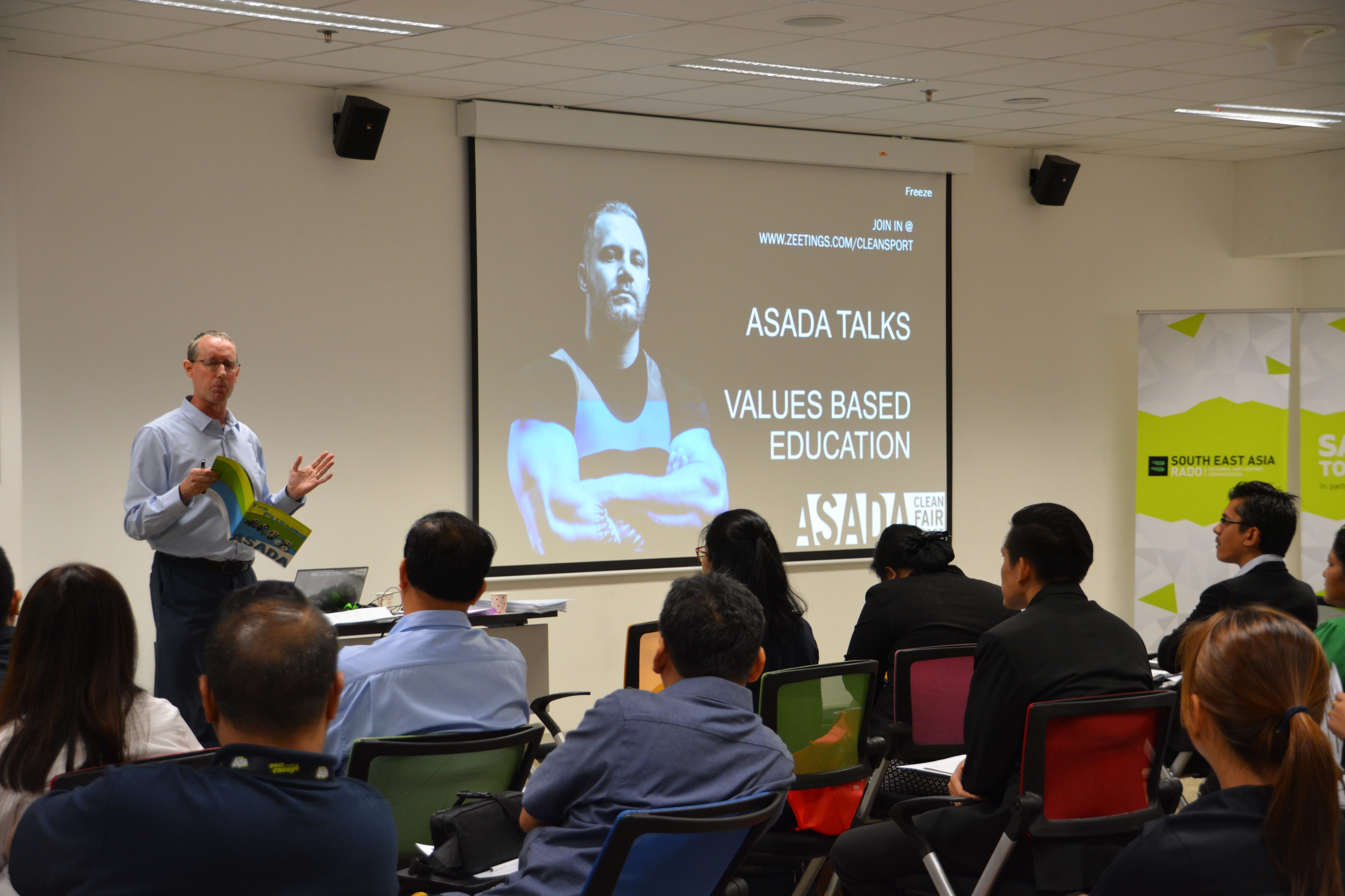 Man speaks to seated audience in classroom; projector slide reads ASADA TALKS: Values Based Education.