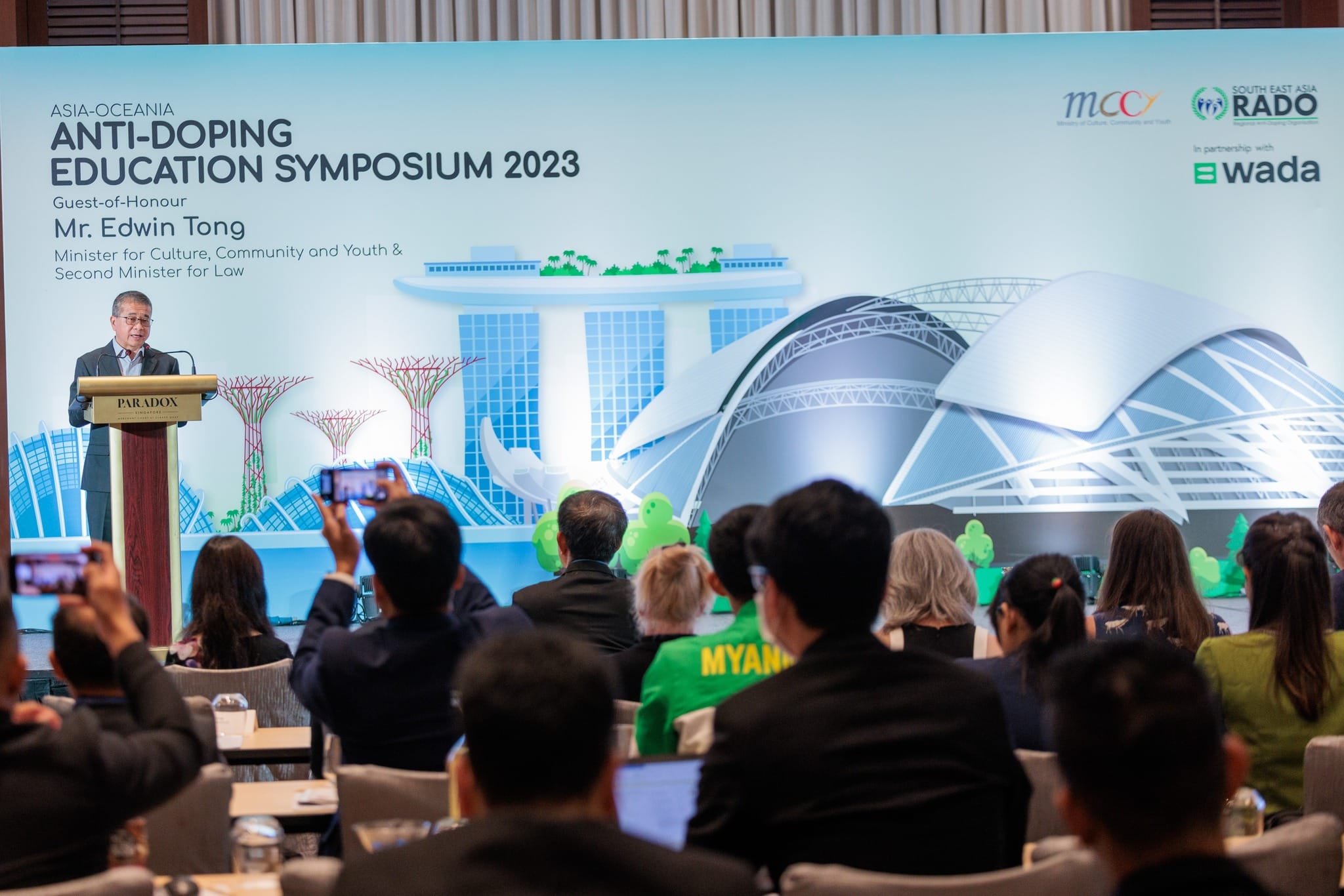 A man speaks at a podium before an audience at the Asia-Oceania Anti-Doping Education Symposium 2023 banner.