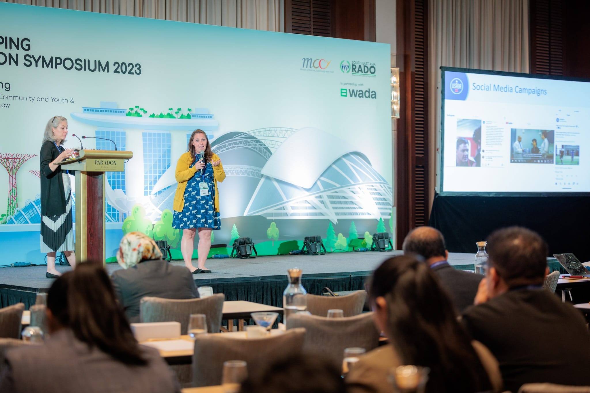 Two women present on stage at the South East Asia RADO Symposium 2023, with an audience seated at tables.