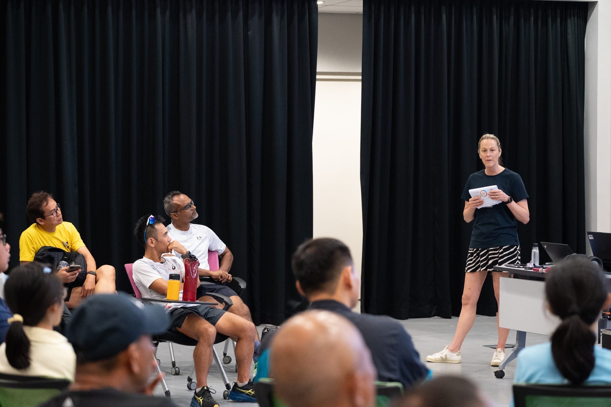 A woman in a black top and striped skirt holds papers while speaking to a seated group in a room with black curtains.