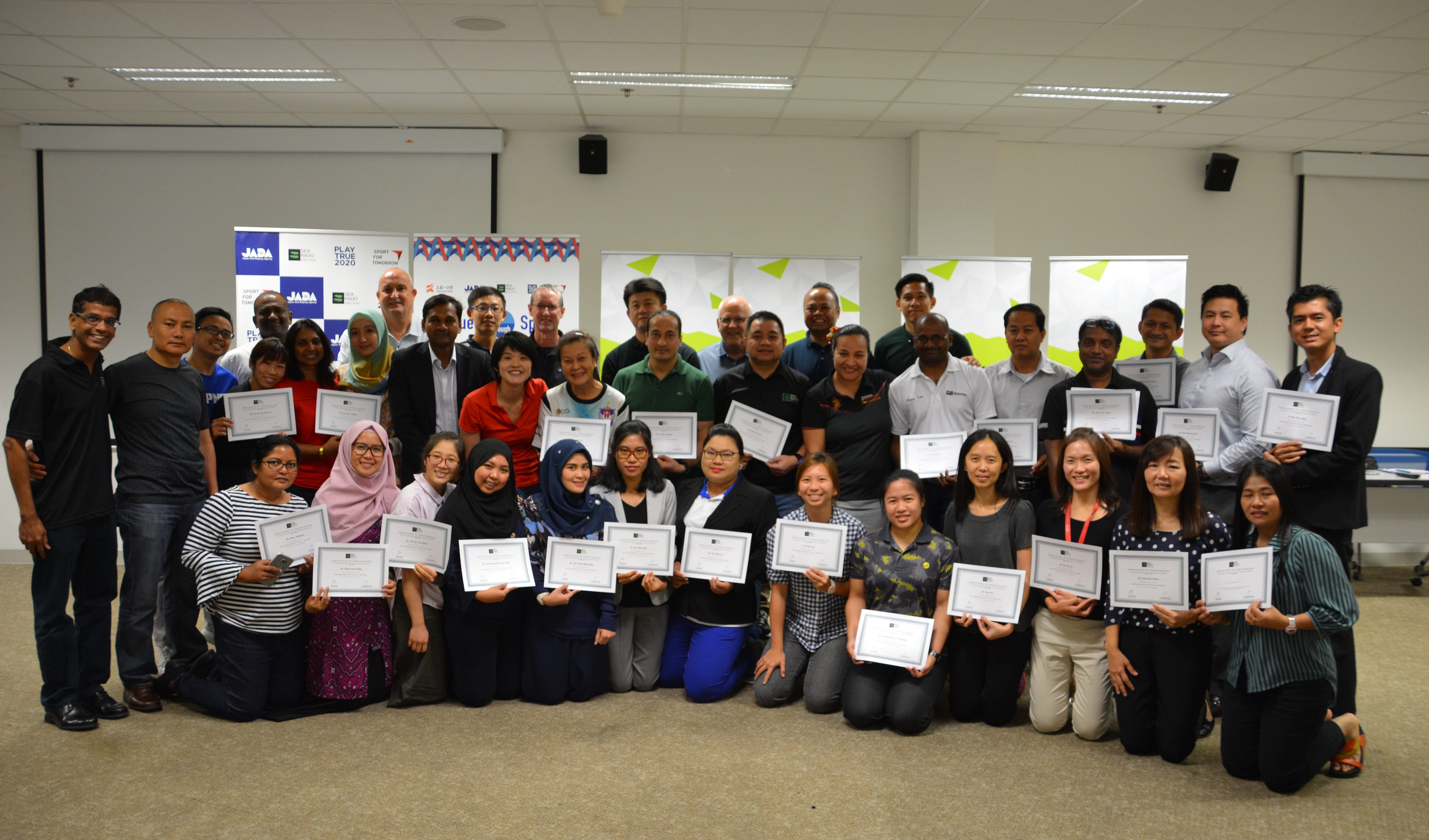 Large group of people posing indoors, most holding certificates, with sponsor banners visible in the background.