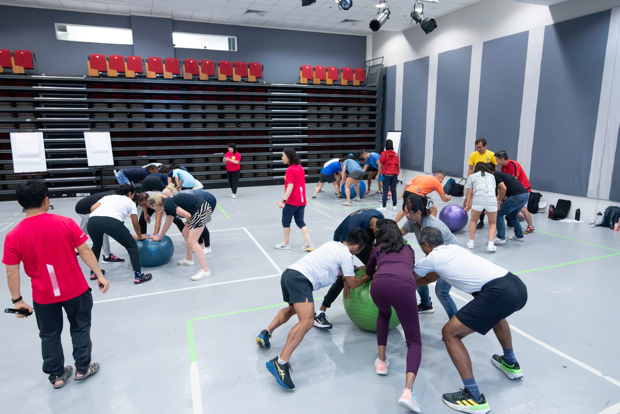 Groups of people in a gym hall doing exercise drills with large fitness balls on a sports court floor.