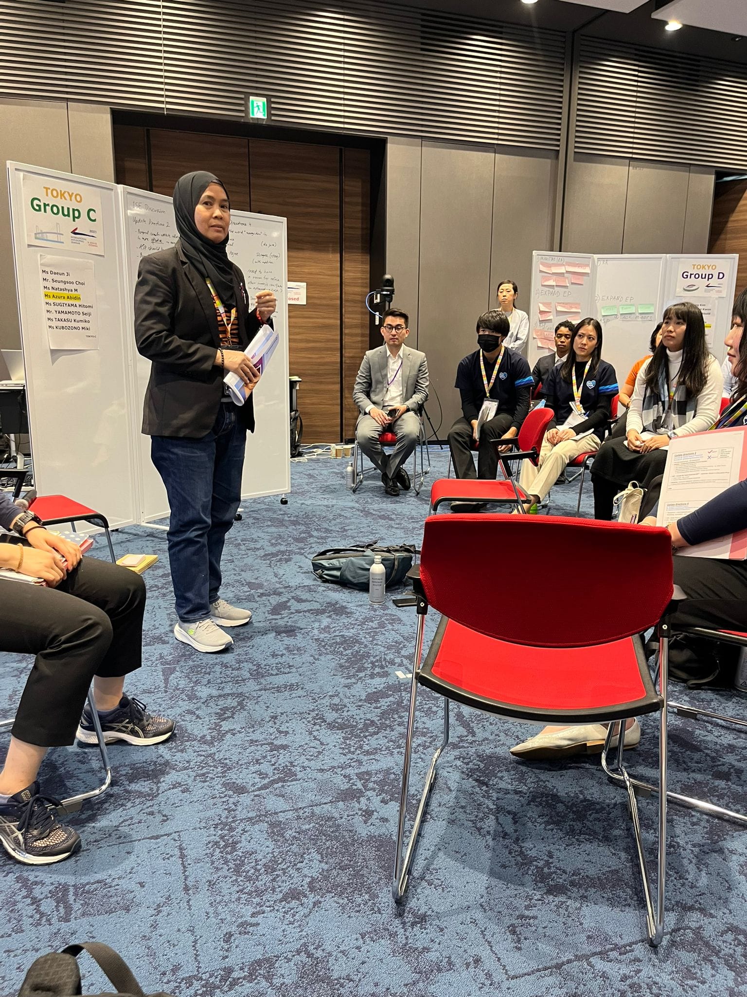 A woman in a hijab stands presenting to a seated group in a workshop room with Tokyo Group C and D boards visible.