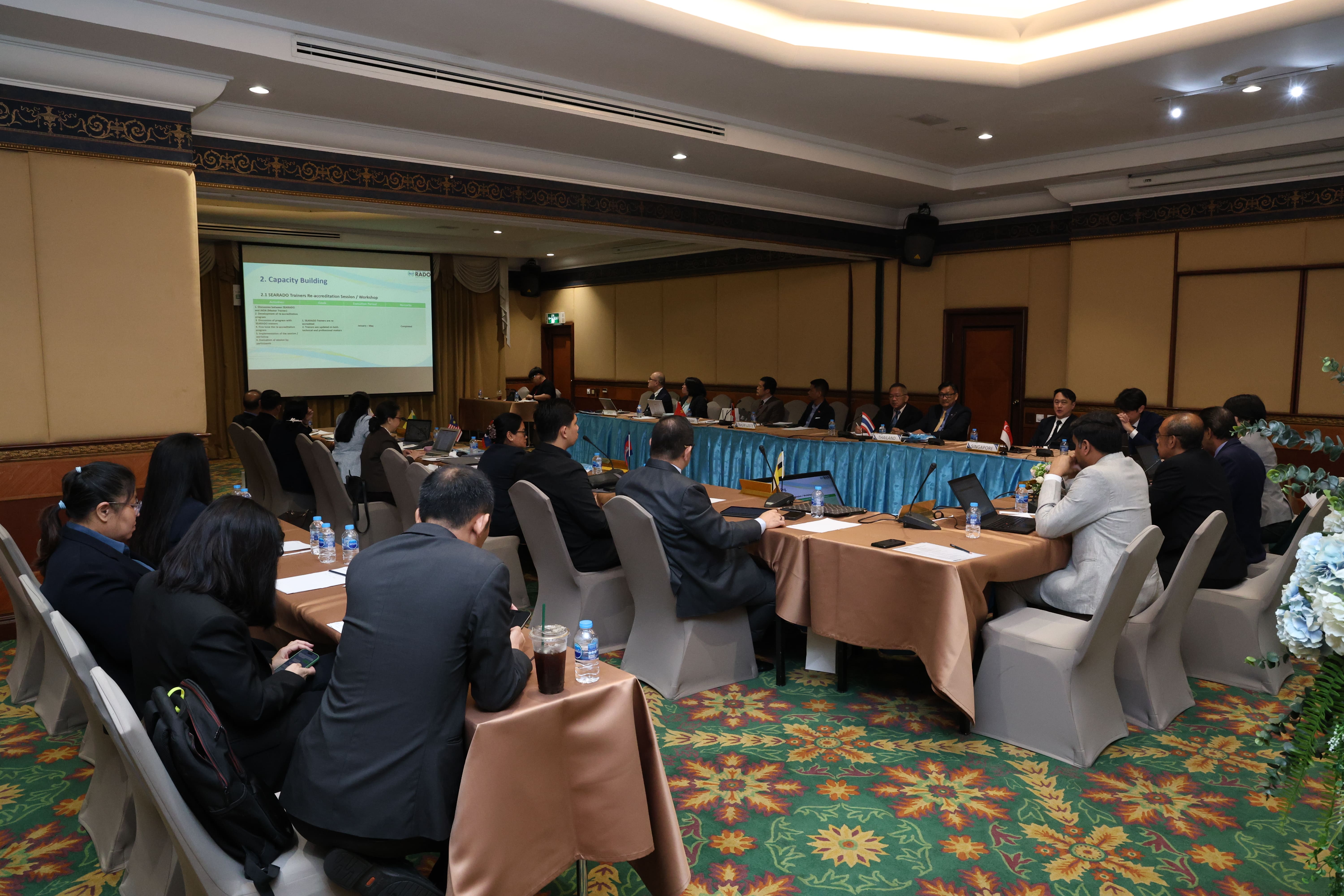 Conference meeting with attendees seated around tables, facing a projected slide at the front.