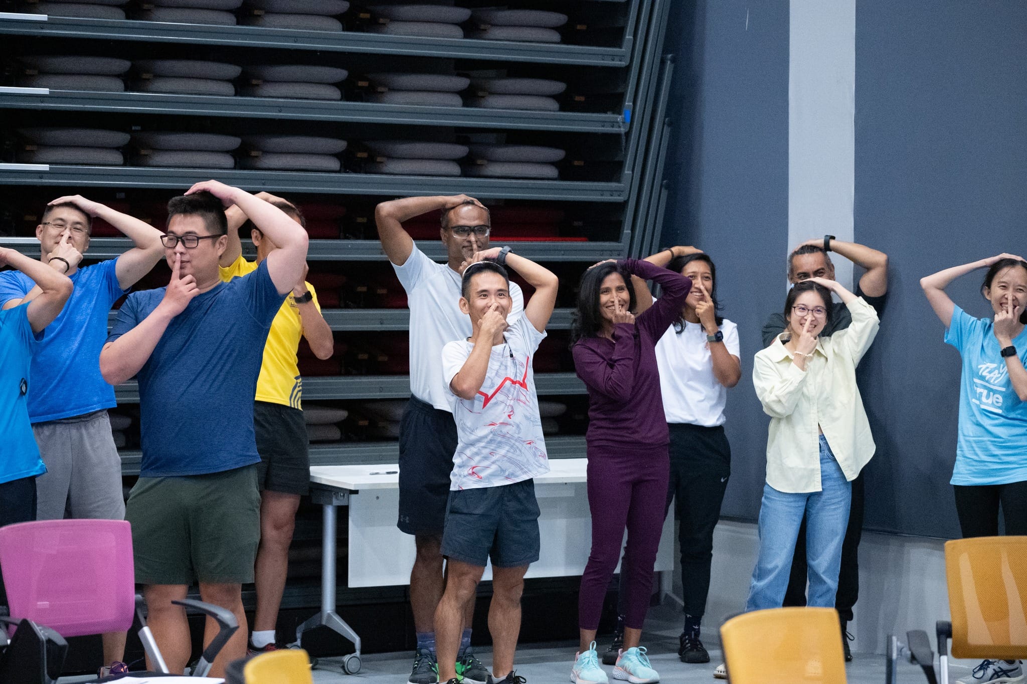 A group of people standing indoors, raising arms over their heads and holding fingers to their lips in unison.