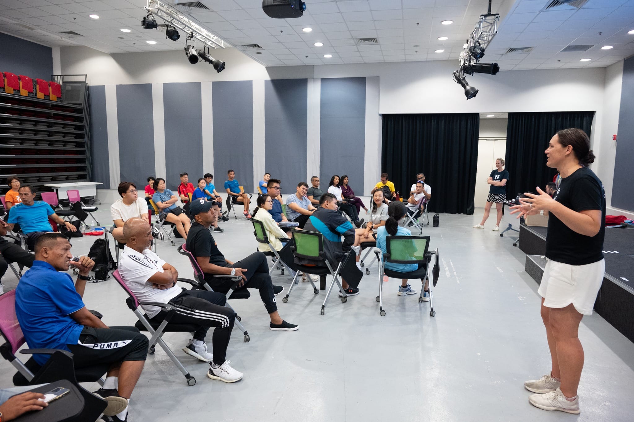 A woman in black shirt and white shorts speaks to a seated group of about 25 people in a large indoor room.