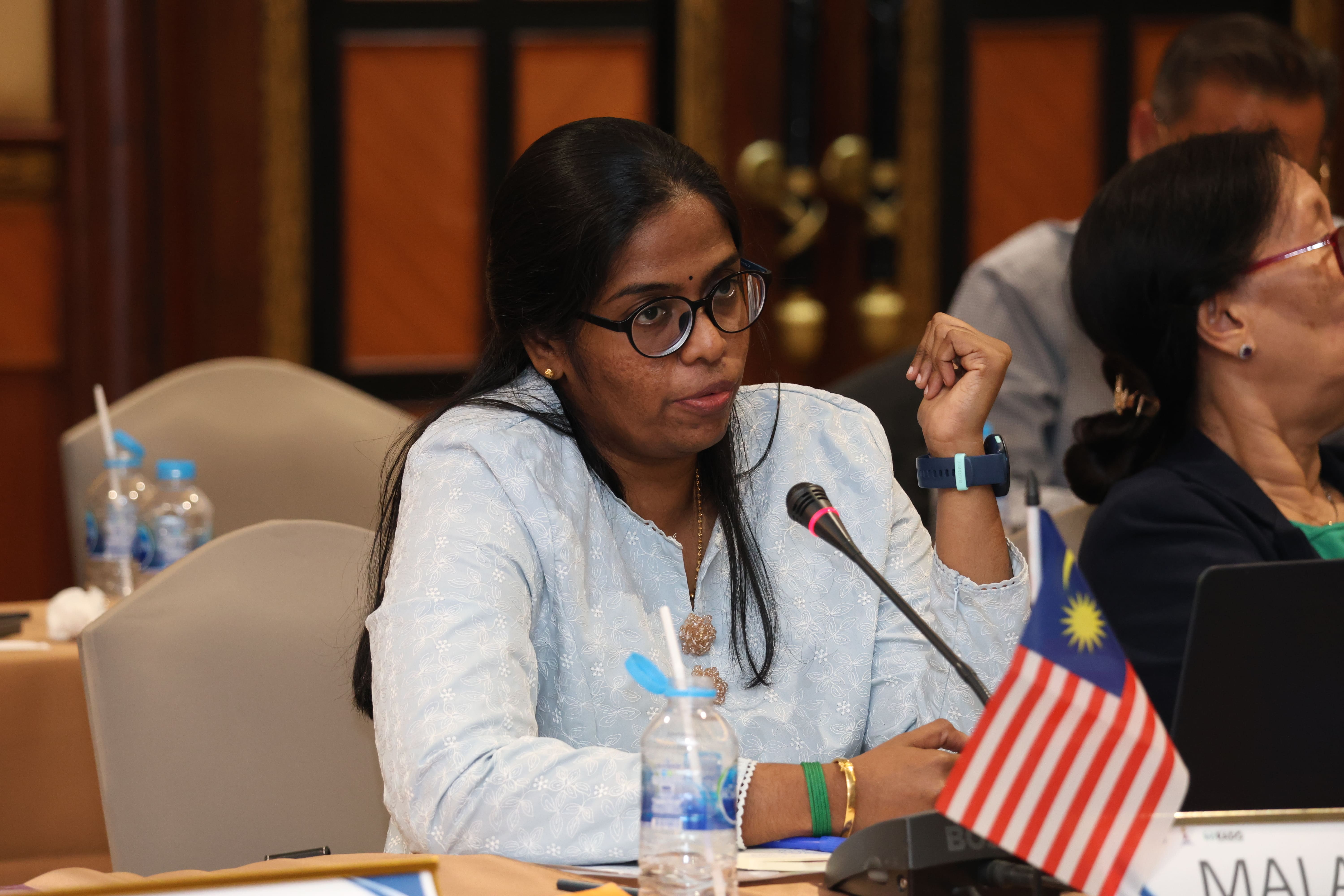 Woman with glasses speaks into a microphone at a meeting table with a Malaysia flag and water bottles.