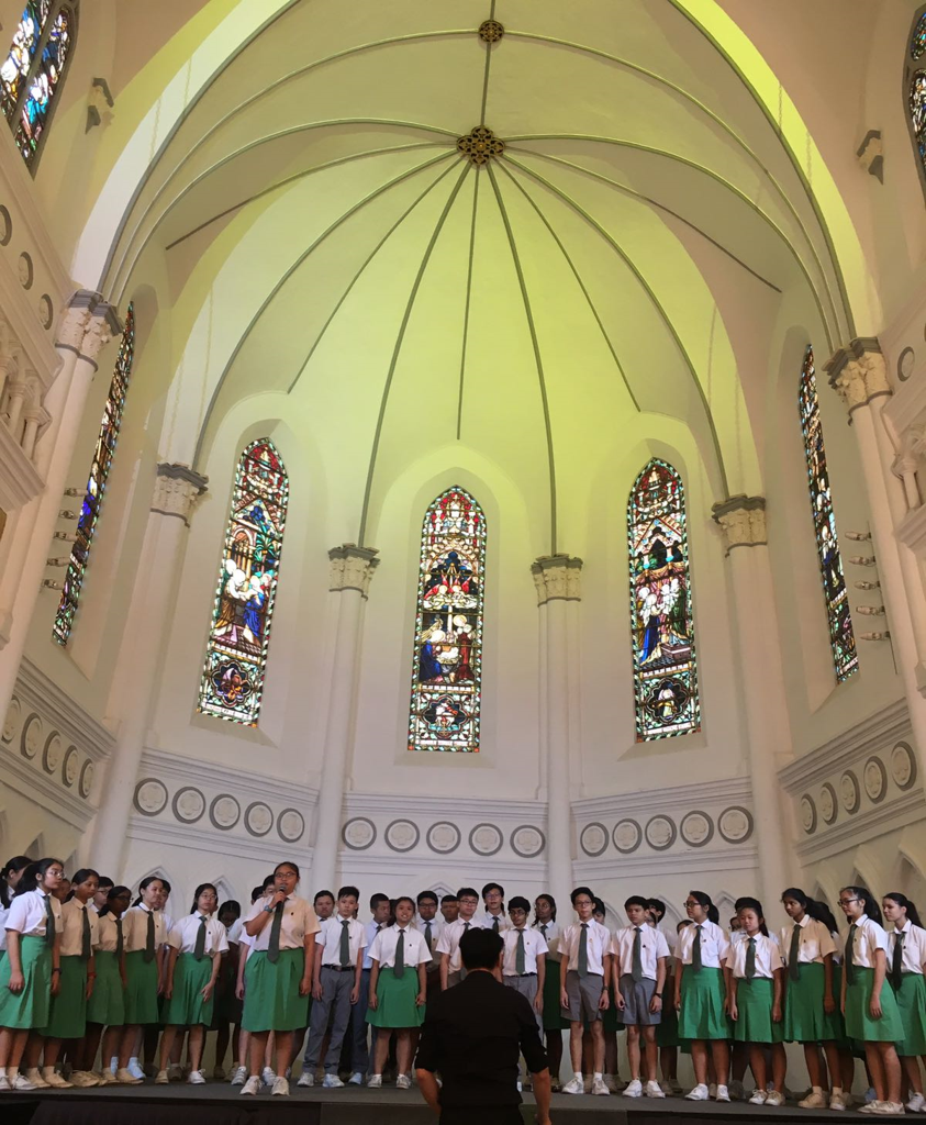 A school choir in white shirts and green uniforms performs inside a church with stained glass windows and a domed ceiling.
