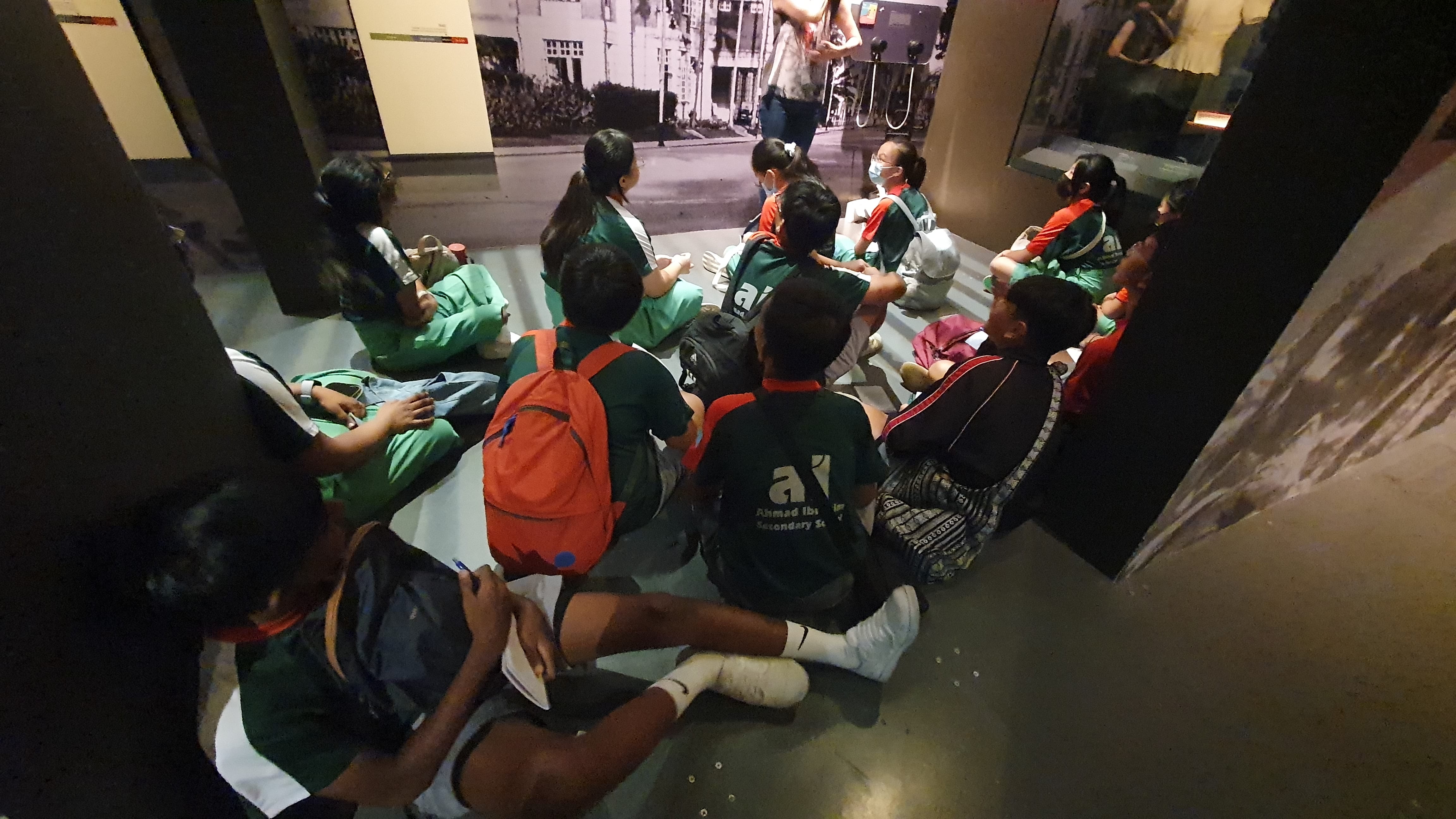 Students in green and dark uniforms sit on a museum floor facing historical photo displays during a school field trip.