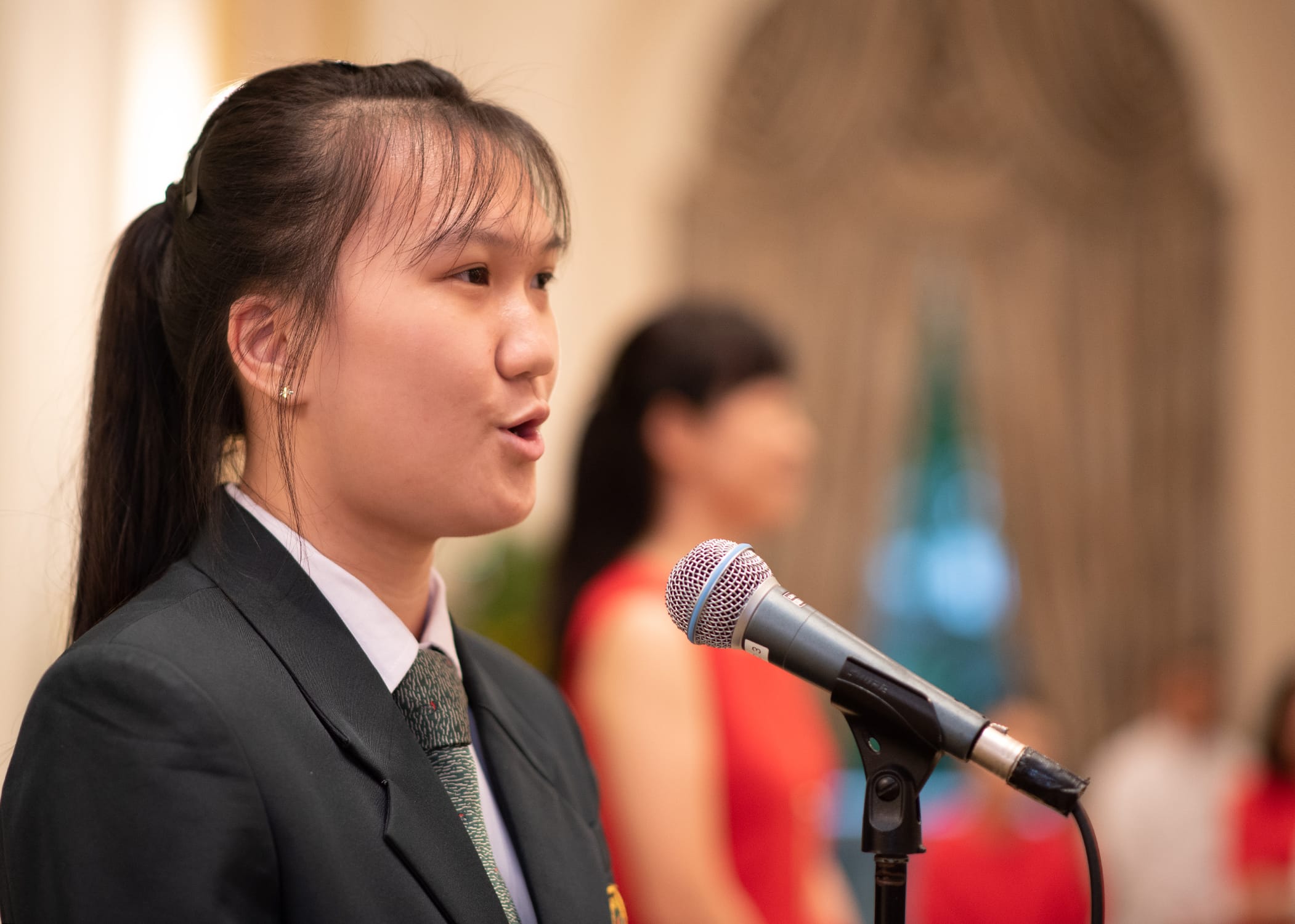 Young woman in a dark blazer and tie speaking into a microphone, with a blurred person in red behind her.