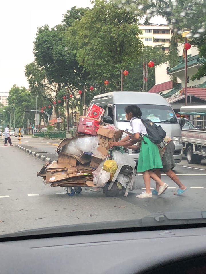 Two people push a cart overloaded with cardboard boxes and bags along a busy street lined with red lanterns.