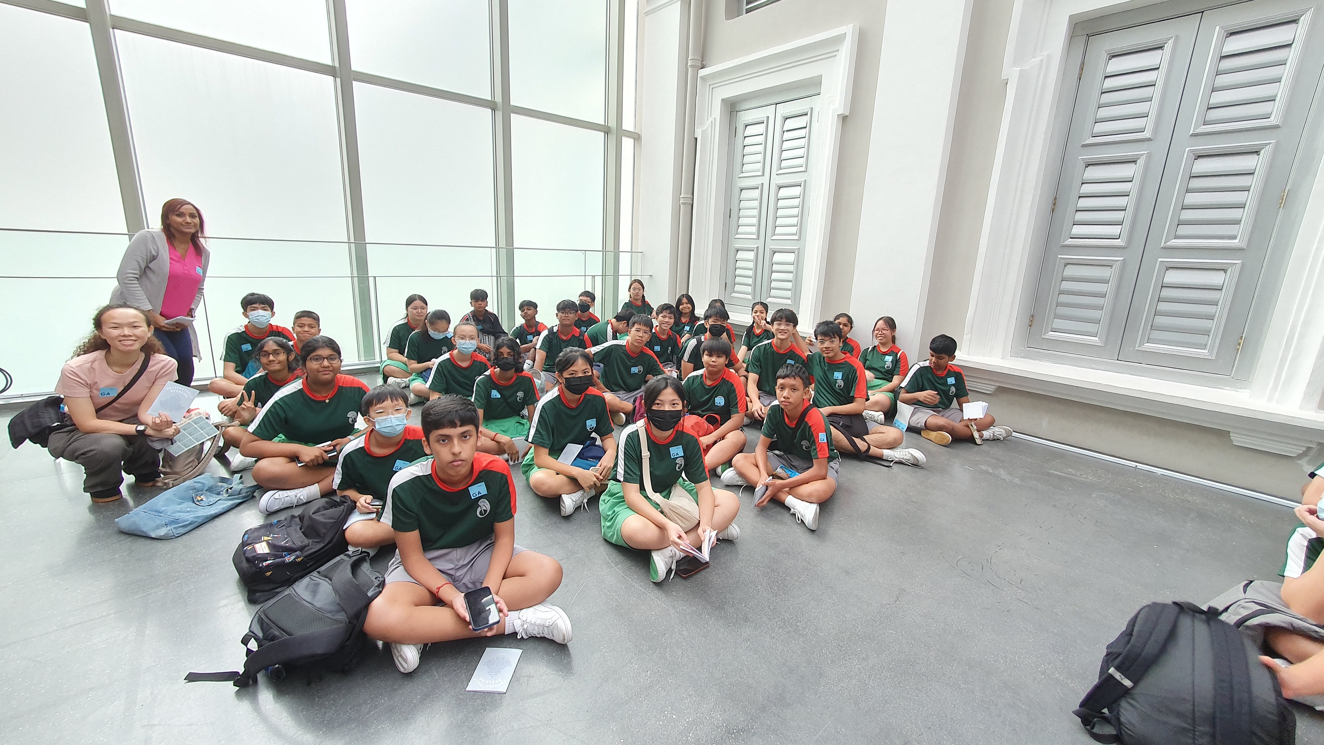 A large group of students in green and red uniforms sit on the floor indoors, accompanied by two adult chaperones.