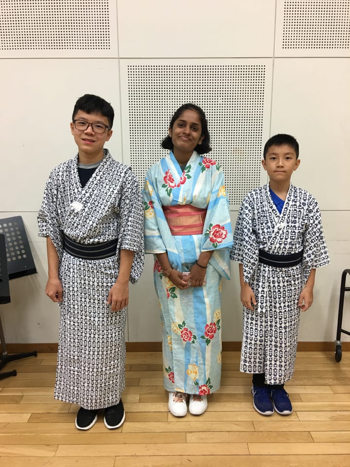Three children wearing traditional Japanese robes stand smiling on a wood floor indoors.