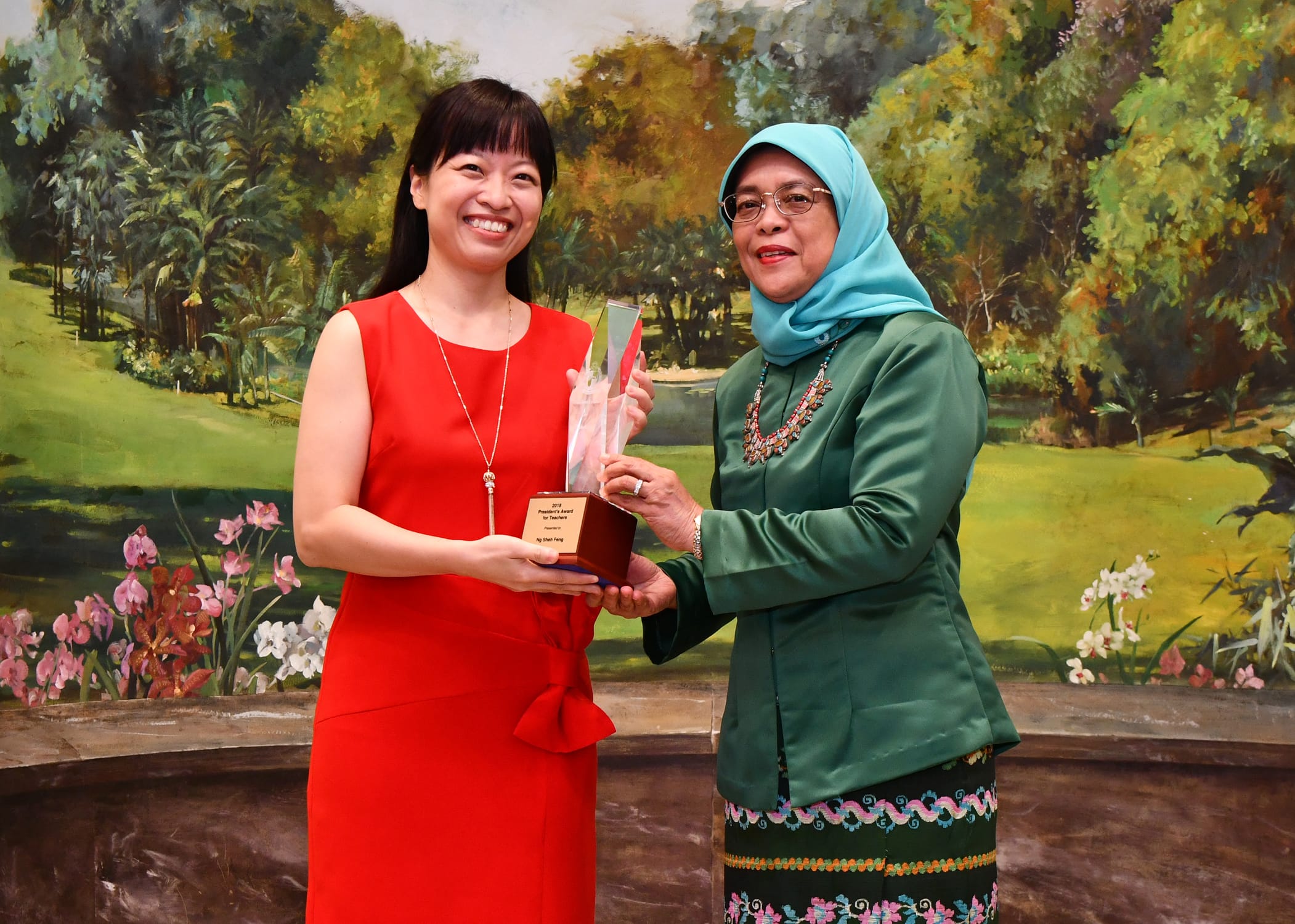 Two women smile while jointly holding a crystal trophy with a wooden base in front of a painted garden backdrop.