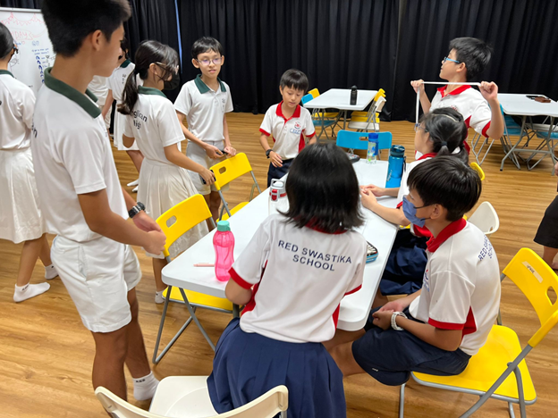 Students in Red Swastika School uniforms gather around tables in a classroom activity session.