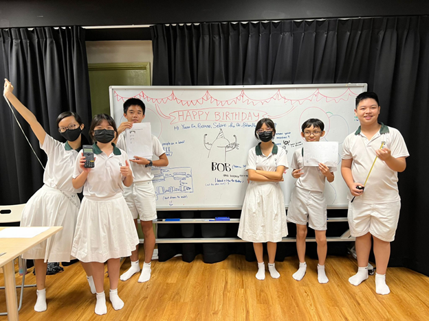 Six students in white uniforms pose in front of a whiteboard with Happy Birthday written on it.
