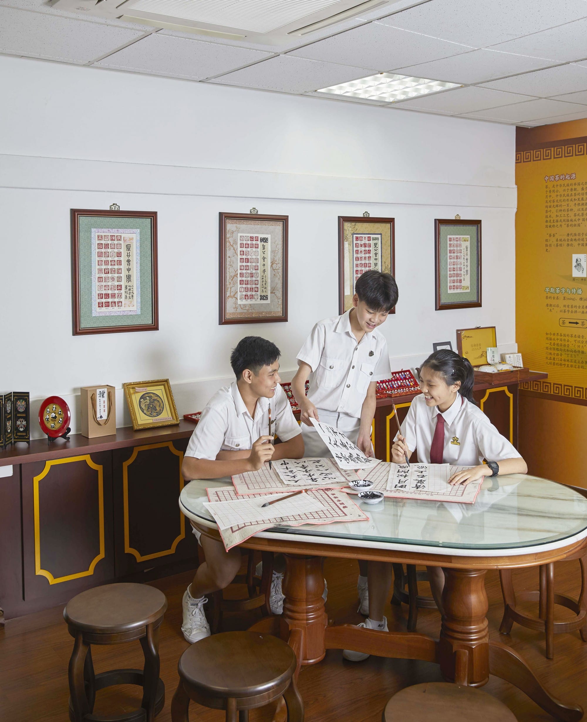 Three students in white uniforms practice Chinese calligraphy at a round table, with framed Chinese artworks on the wall.