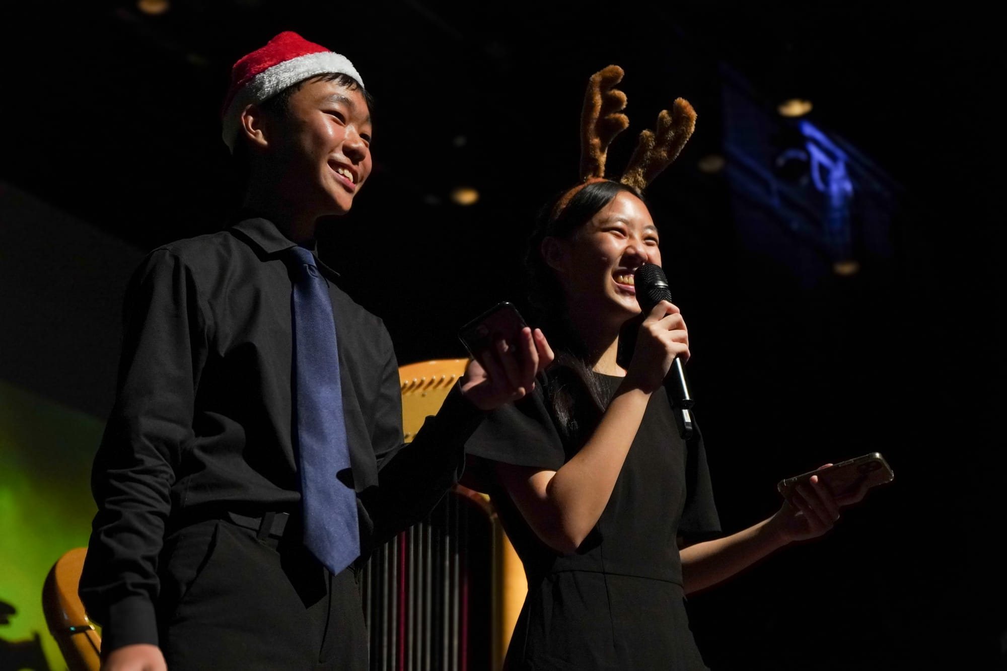 Two smiling teens on stage, one in a Santa hat and one with reindeer antlers, holding a microphone and phone.