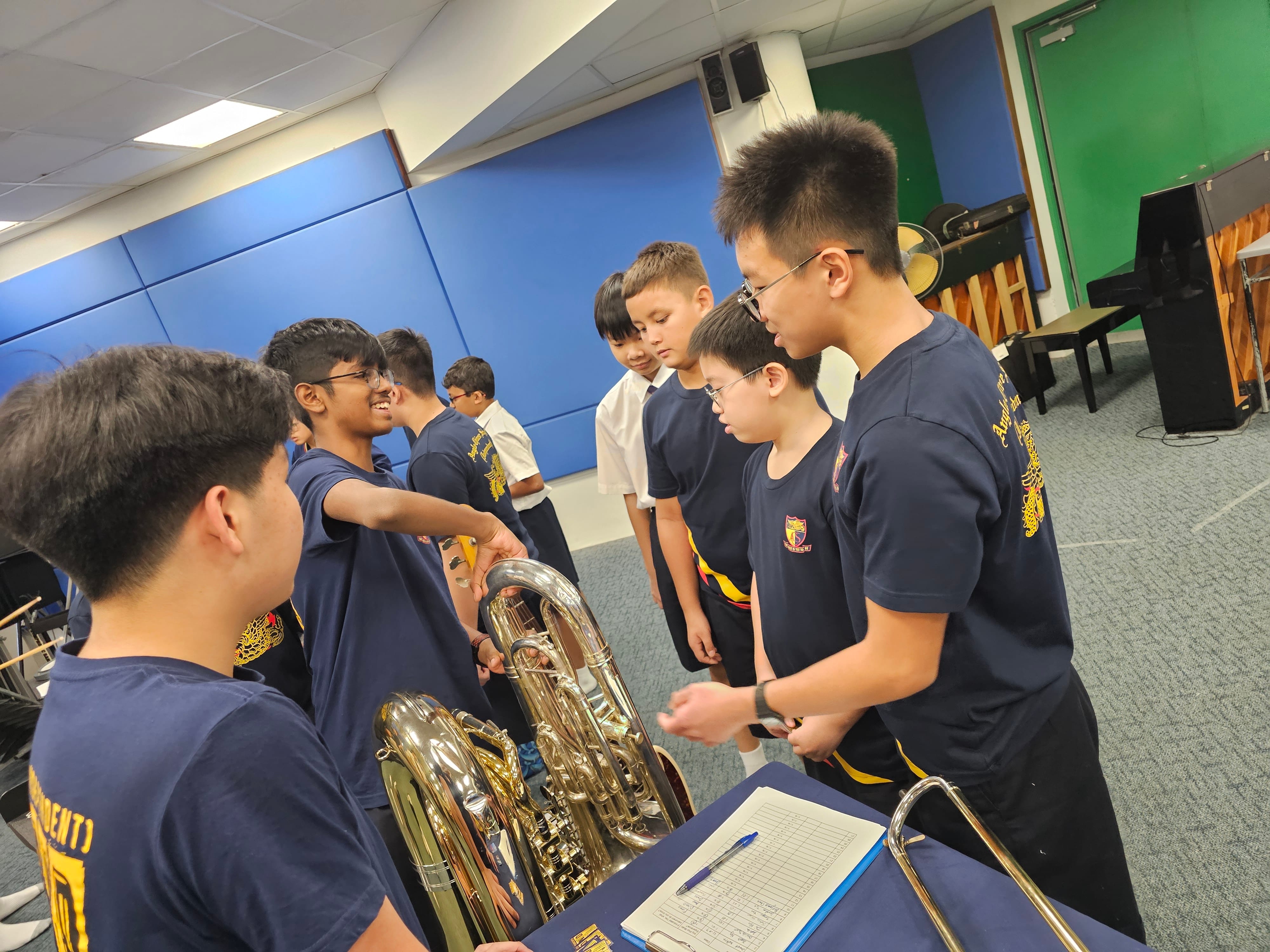 Students in navy uniforms gather around a brass tuba in a music room with a piano and sheet music nearby.