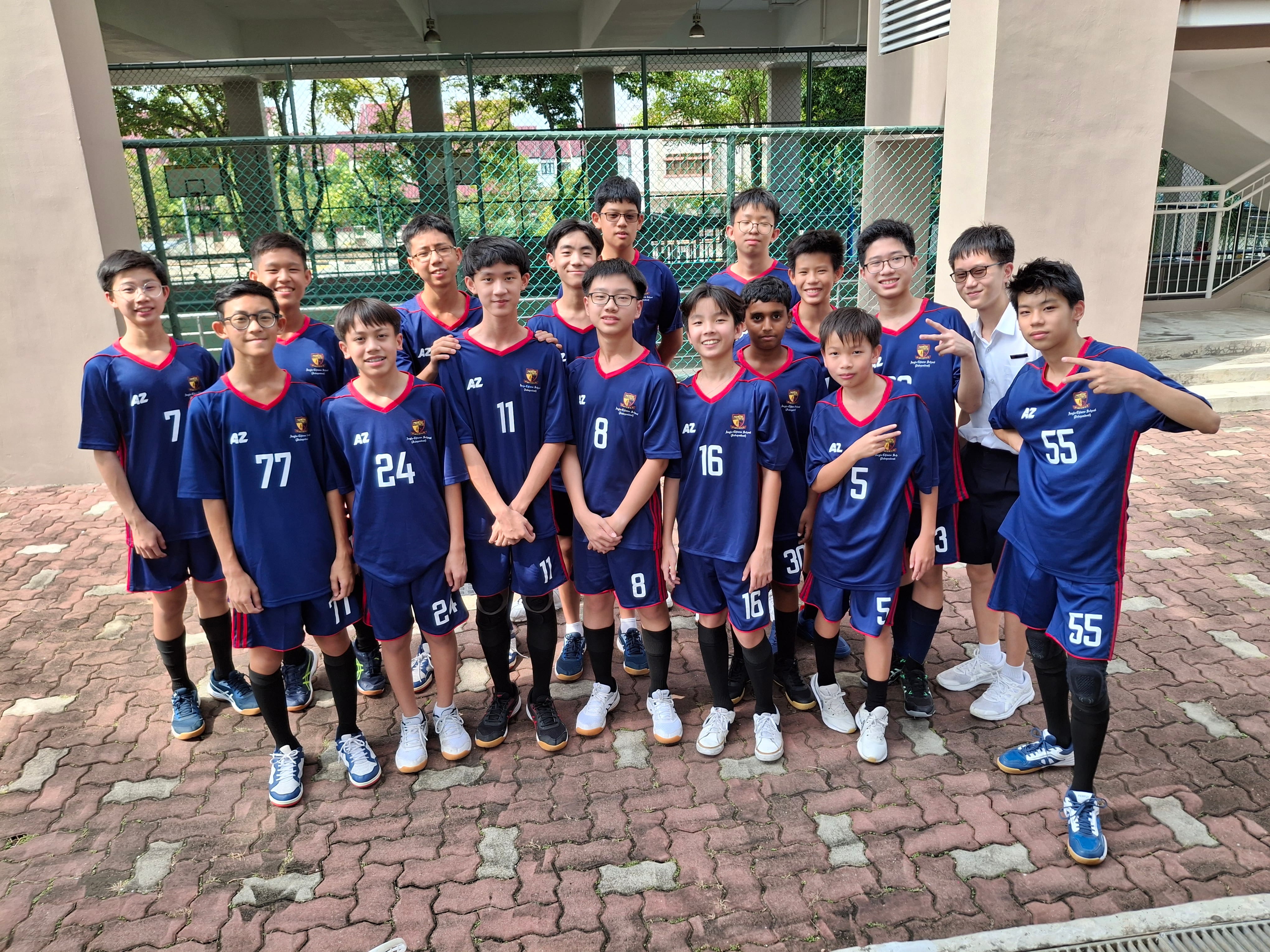 Group of boys in blue jerseys pose outdoors by a chain-link fence, some flashing peace signs.