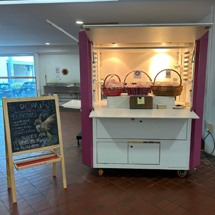 A pink and white donut kiosk on wheels with baskets of donuts and a chalkboard menu sign beside it.