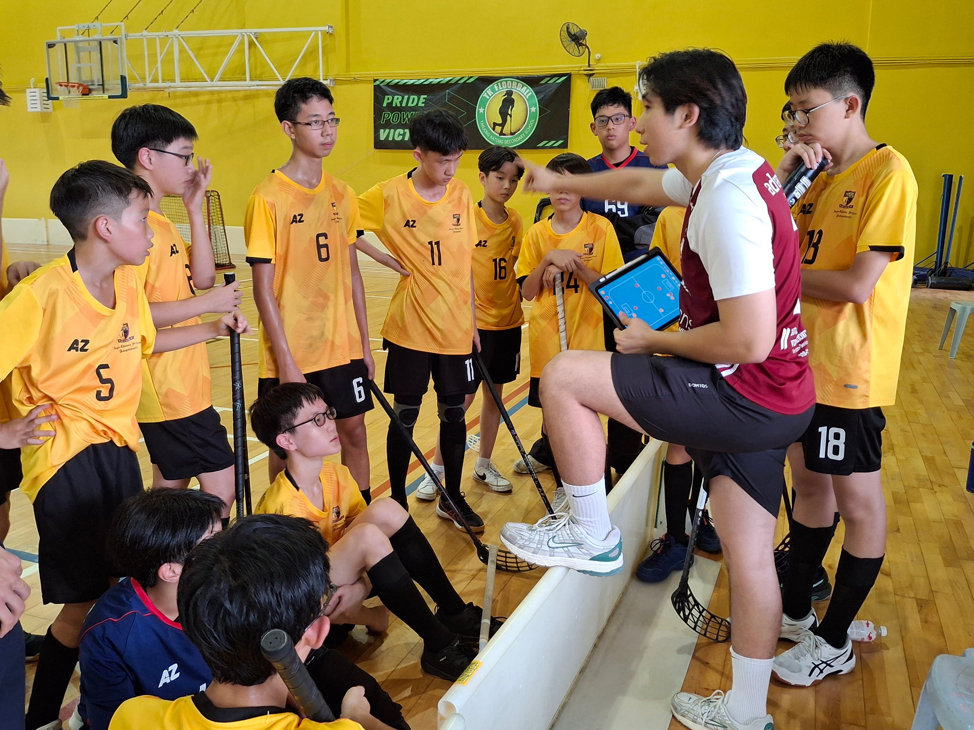 A coach holding a tablet gives instructions to young floorball players in yellow jerseys inside a gymnasium.