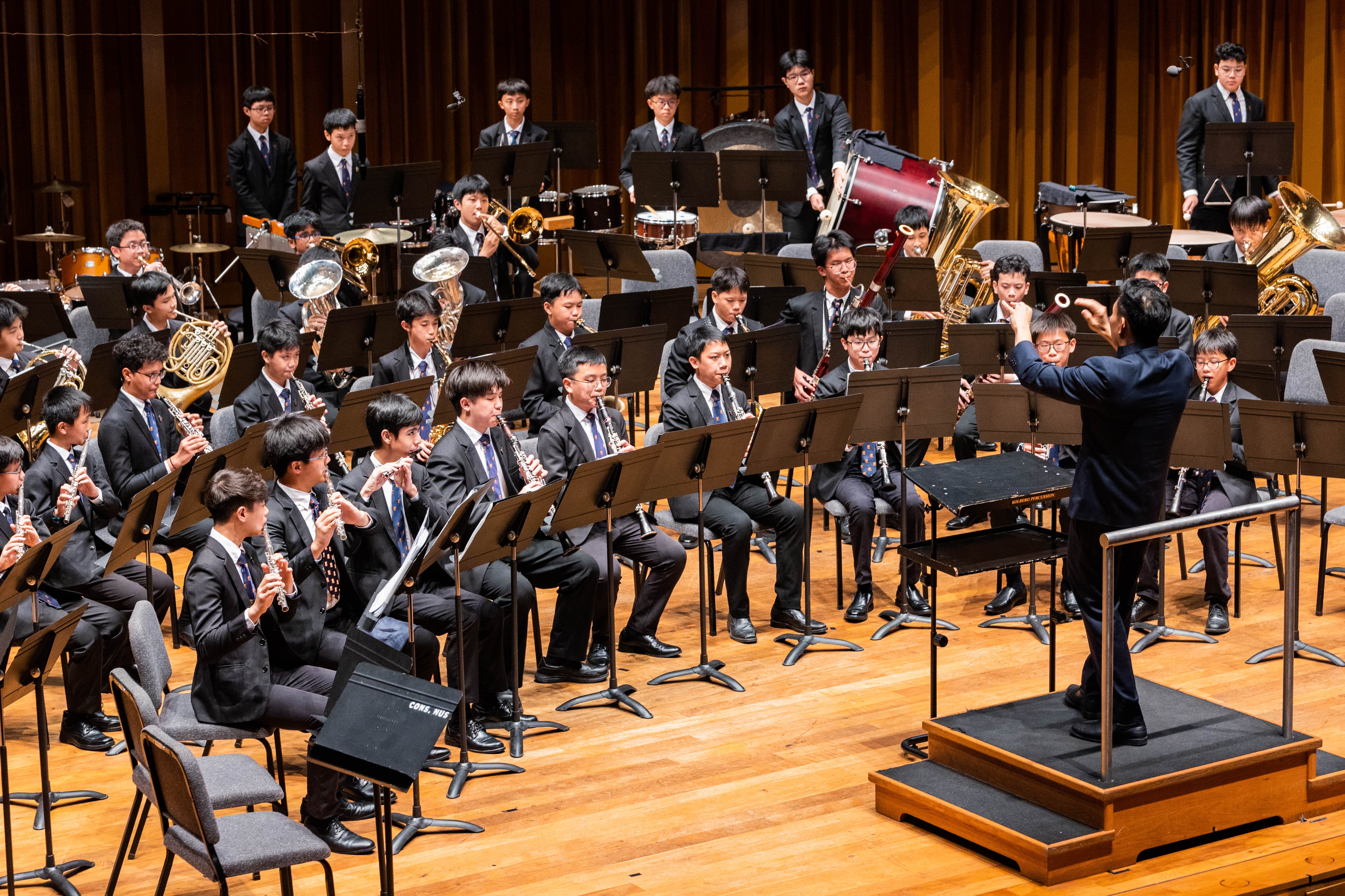 A conductor leads a large youth wind band in formal black suits performing on a wooden concert stage.