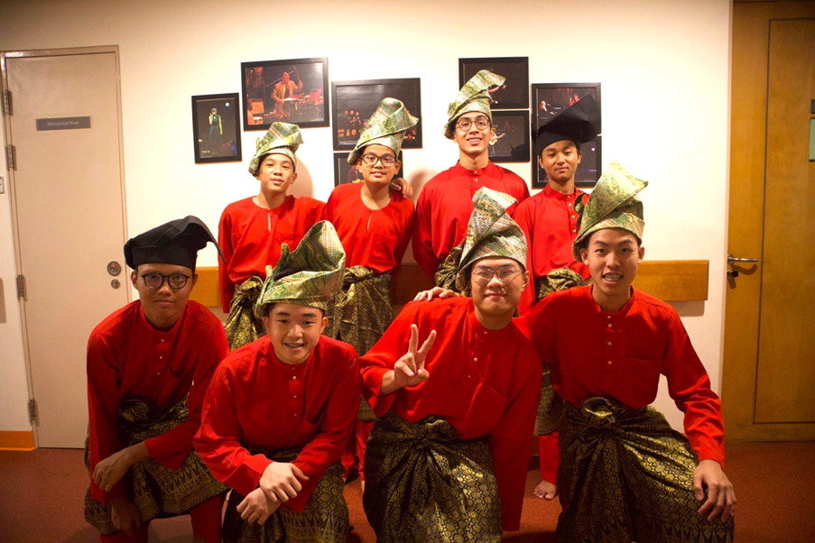 Seven young men in red traditional Malay baju melayu with gold sarongs and decorative headwear pose indoors.