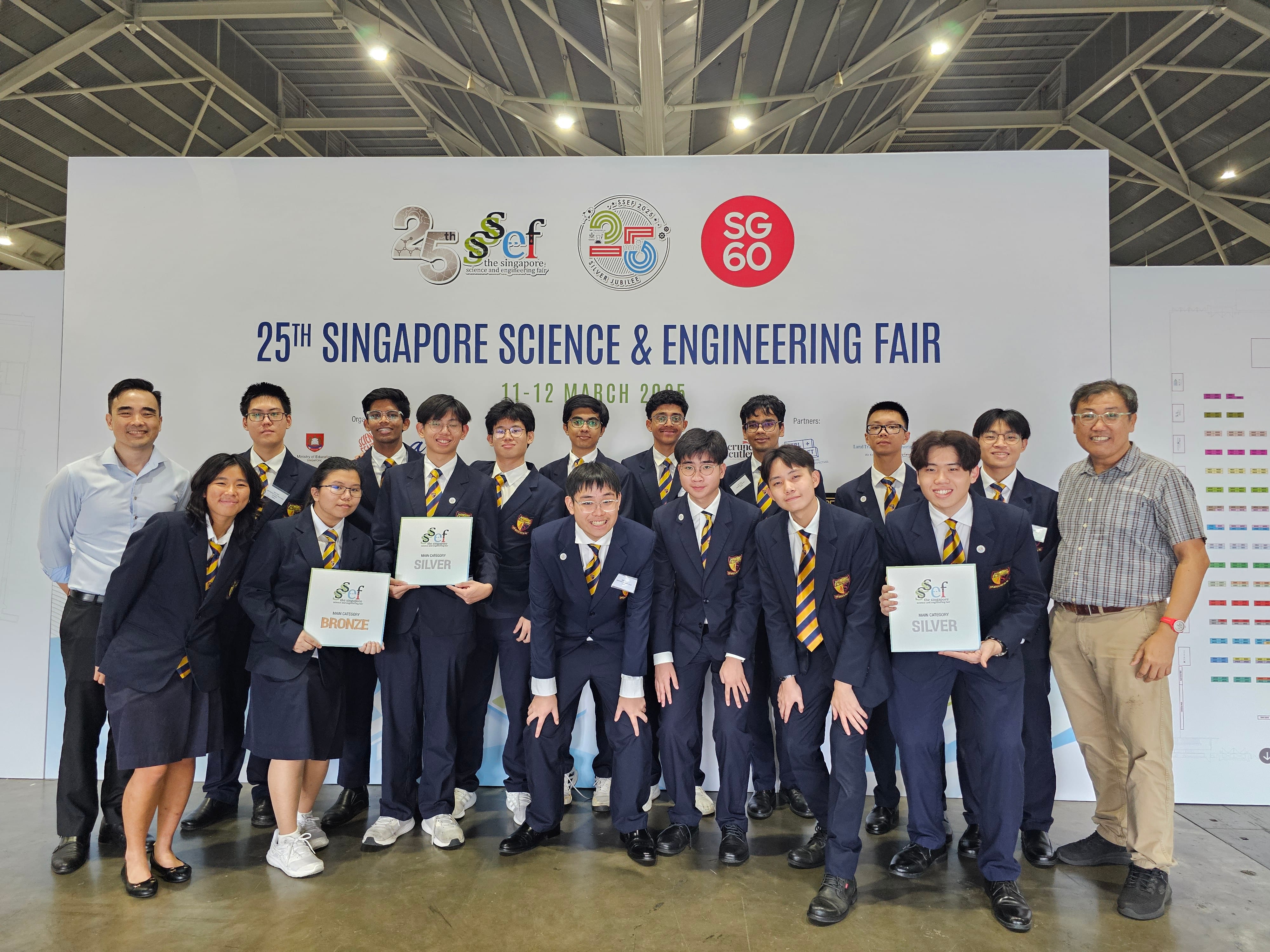 A group of students with navy blue blazers over their school uniforms pose at the 25th Singapore Science & Engineering Fair. Three hold certificates. The mood is celebratory.