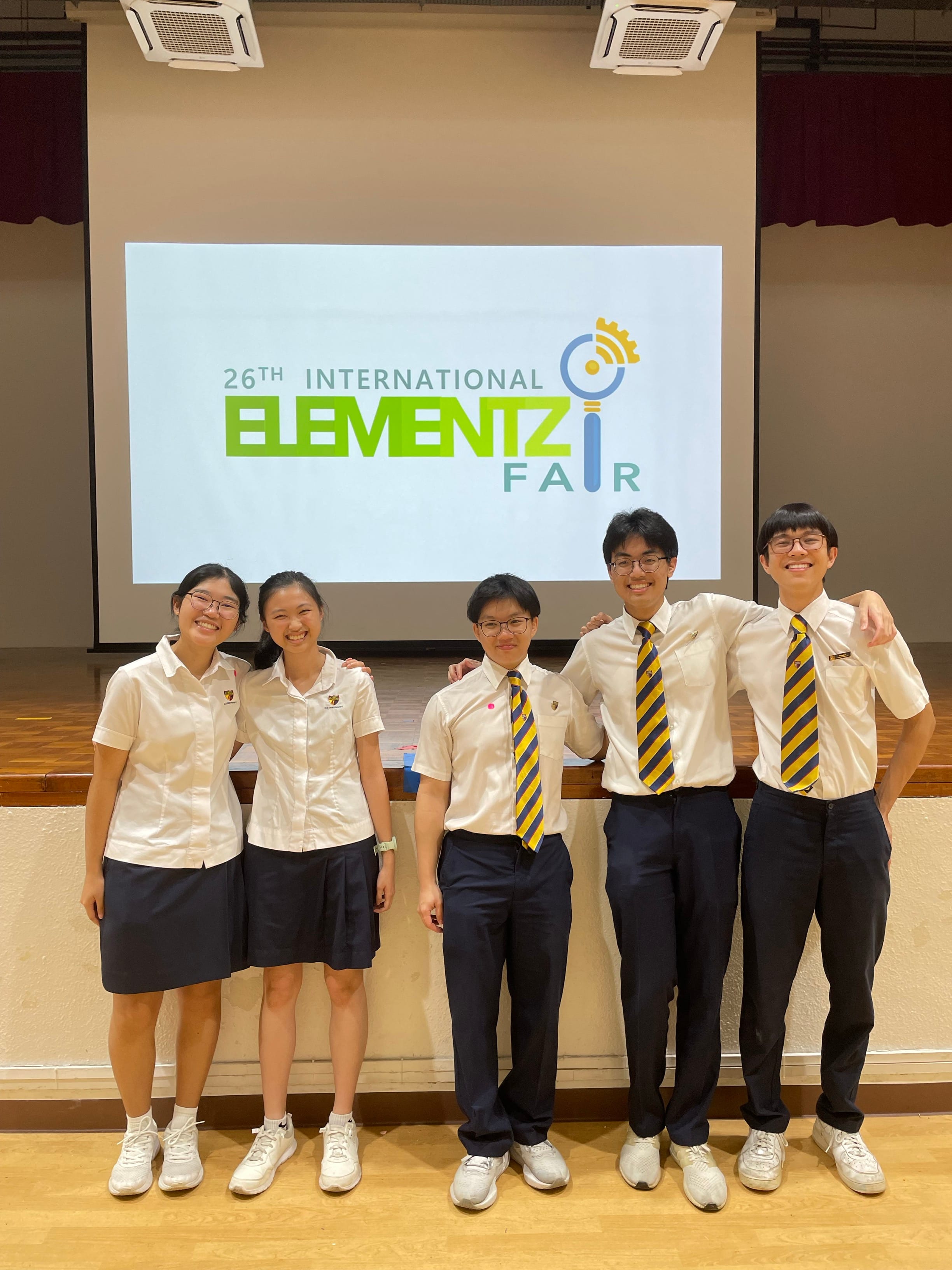 Five students in school uniform smile in front of a stage. Behind them, there is a screen displaying "26th International Elementz Fair". They convey a sense of achievement and camaraderie.