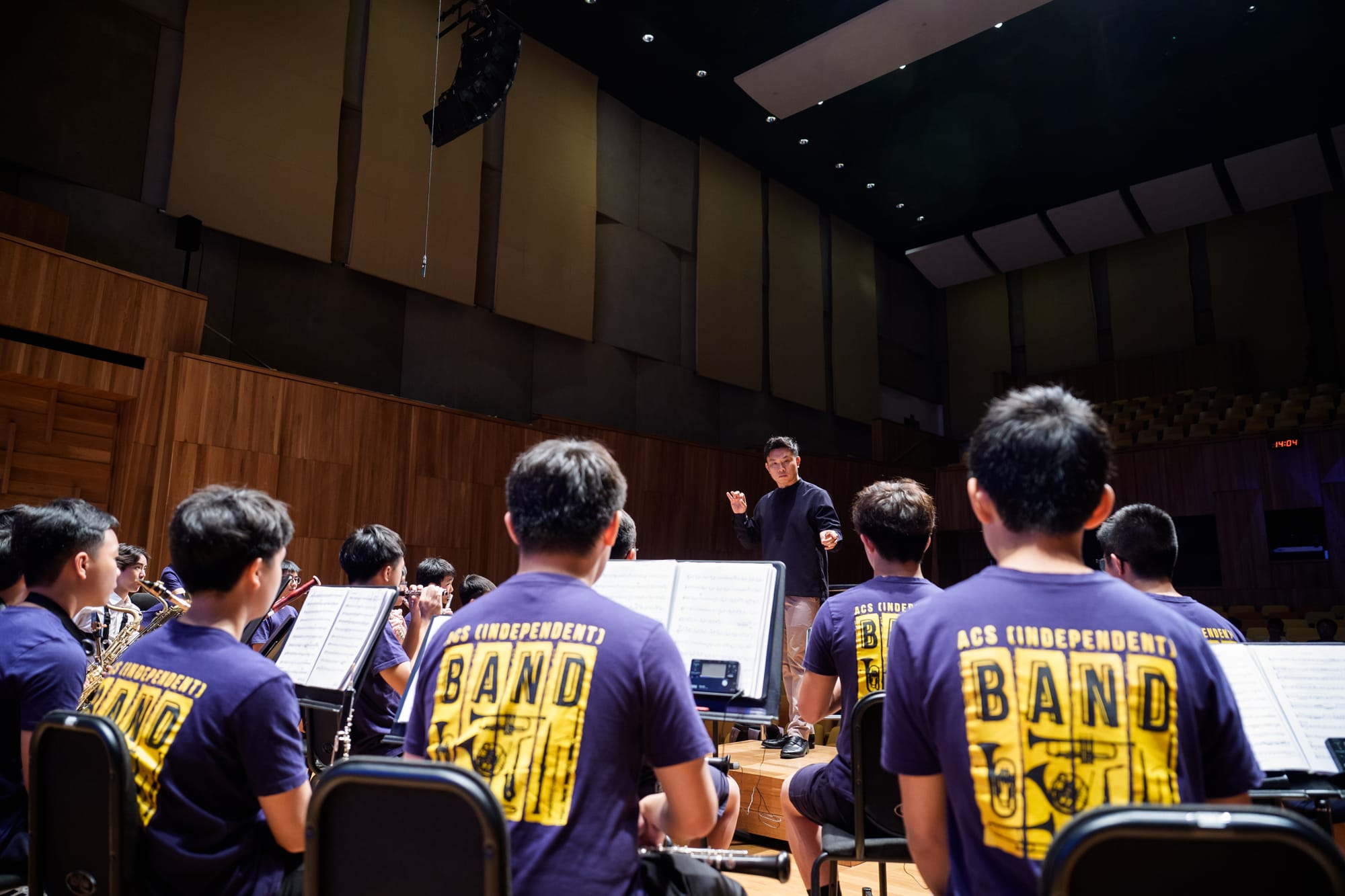 A conductor leads ACS Independent Band students in purple shirts during a rehearsal in a concert hall.