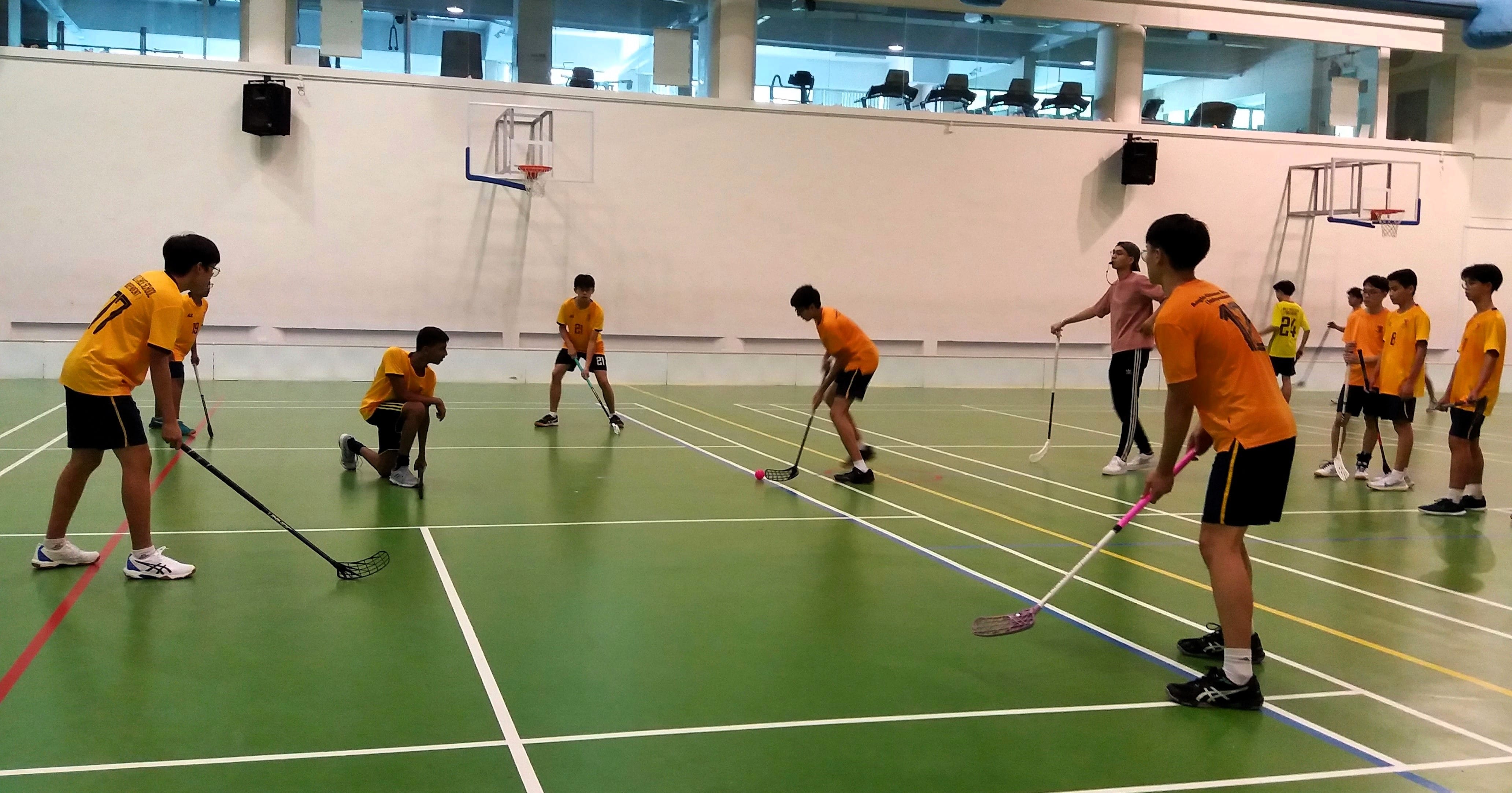 Students in orange jerseys play floorball with sticks on a green indoor court with basketball hoops visible.