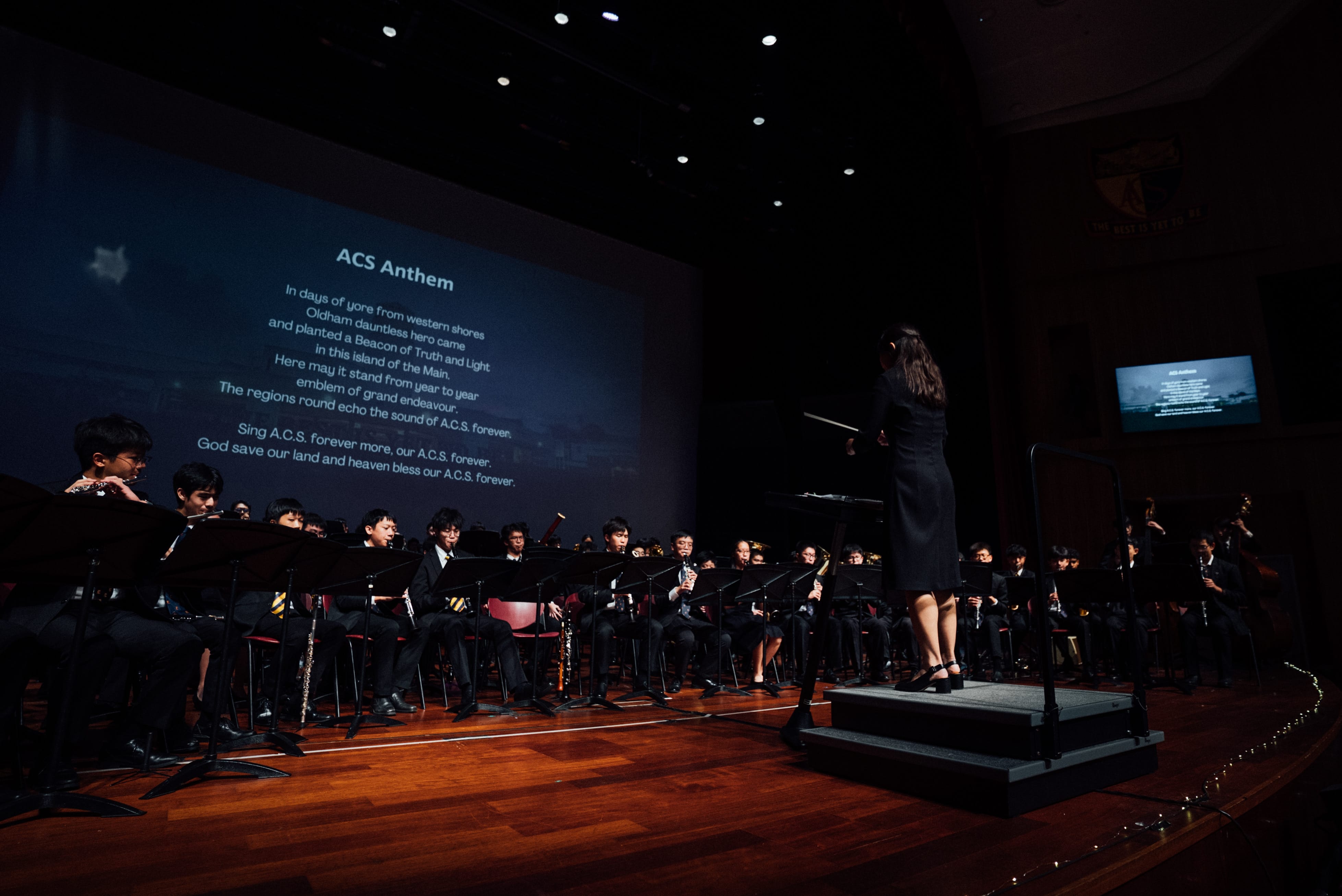 A conductor leads a youth orchestra on a dark stage with the ACS Anthem lyrics displayed on a large screen behind them.
