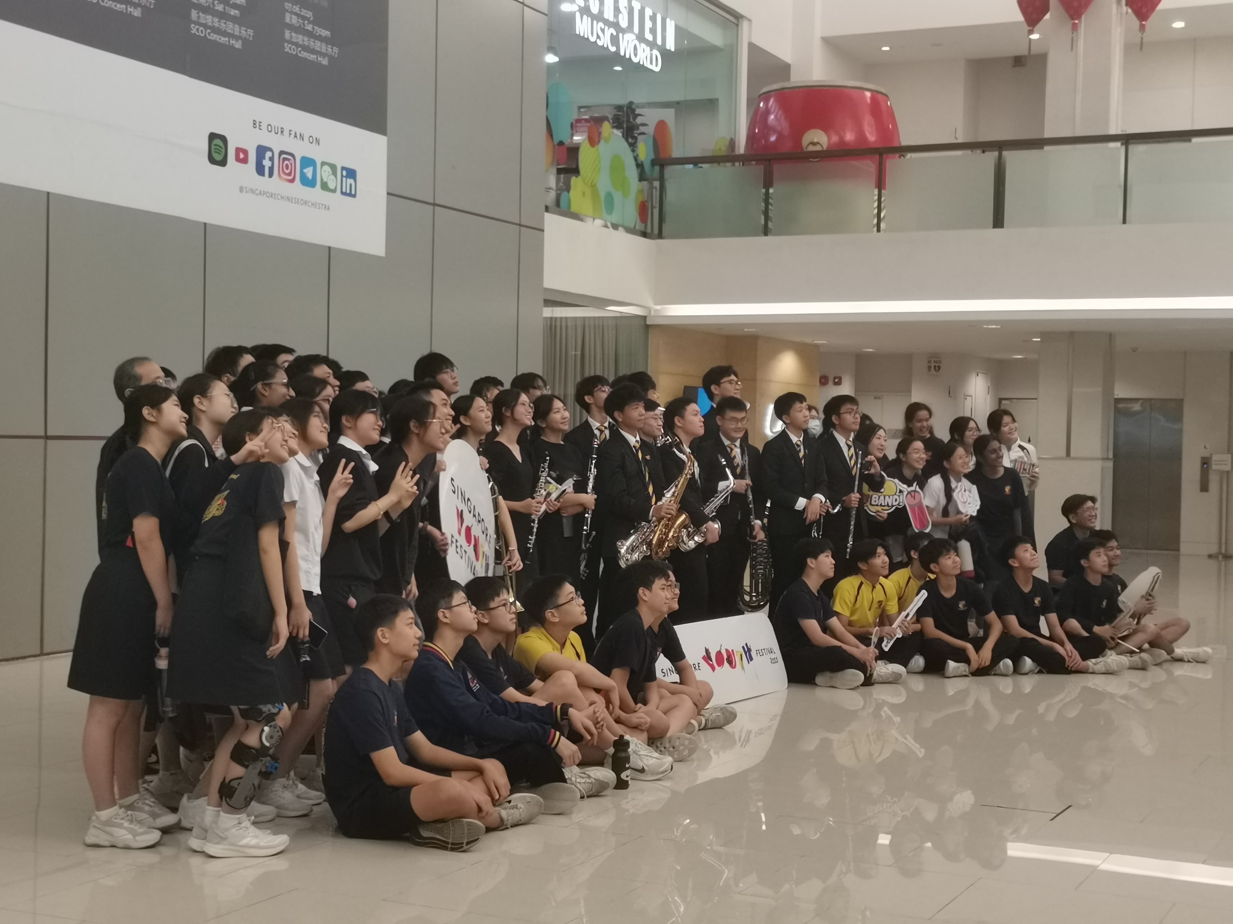 A large group of students with musical instruments pose for a photo in a mall lobby near a Singapore Youth Festival sign.