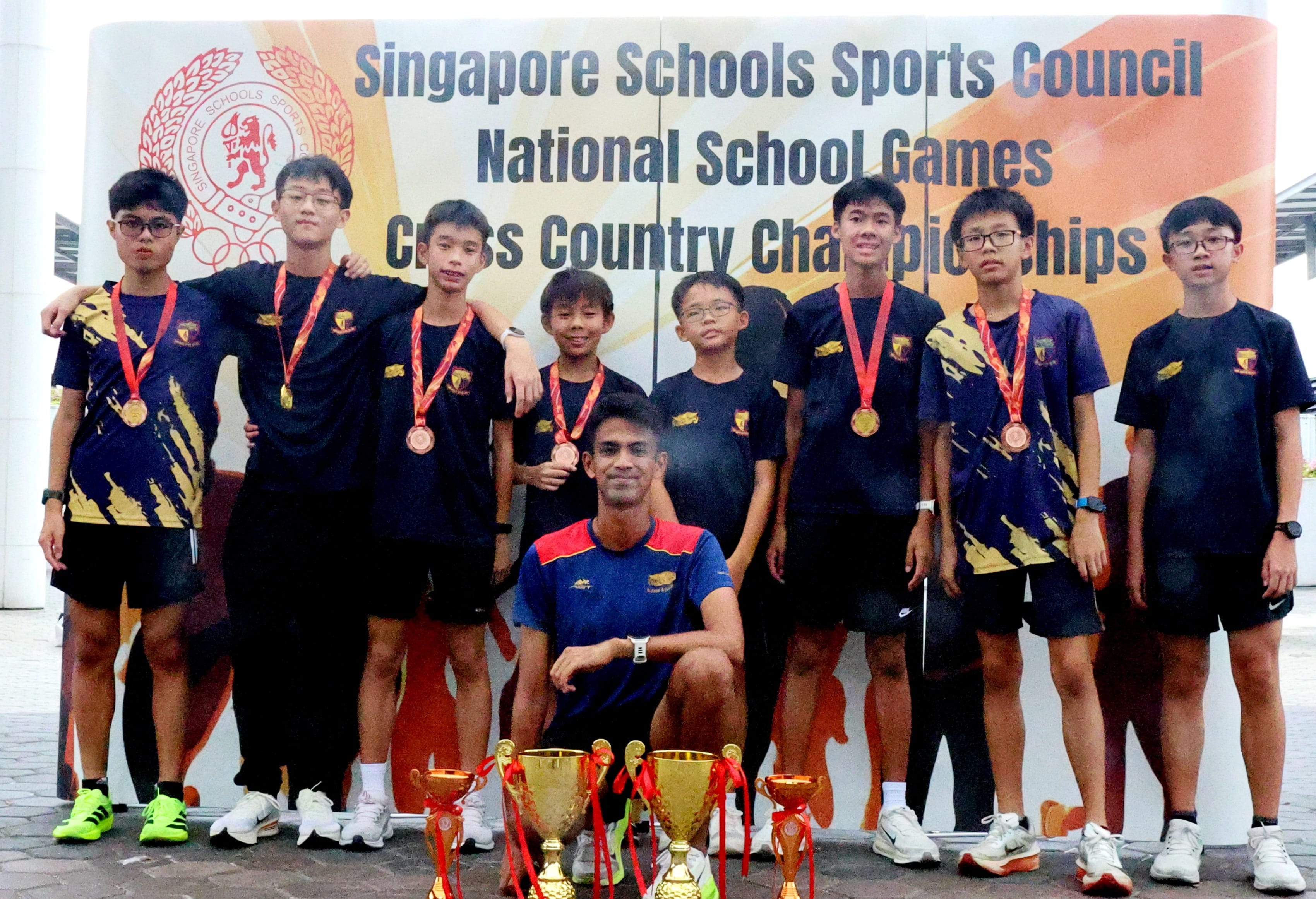 Eight boys with medals and trophies pose before a Singapore Schools Sports Council National School Games Cross Country Championships banner.
