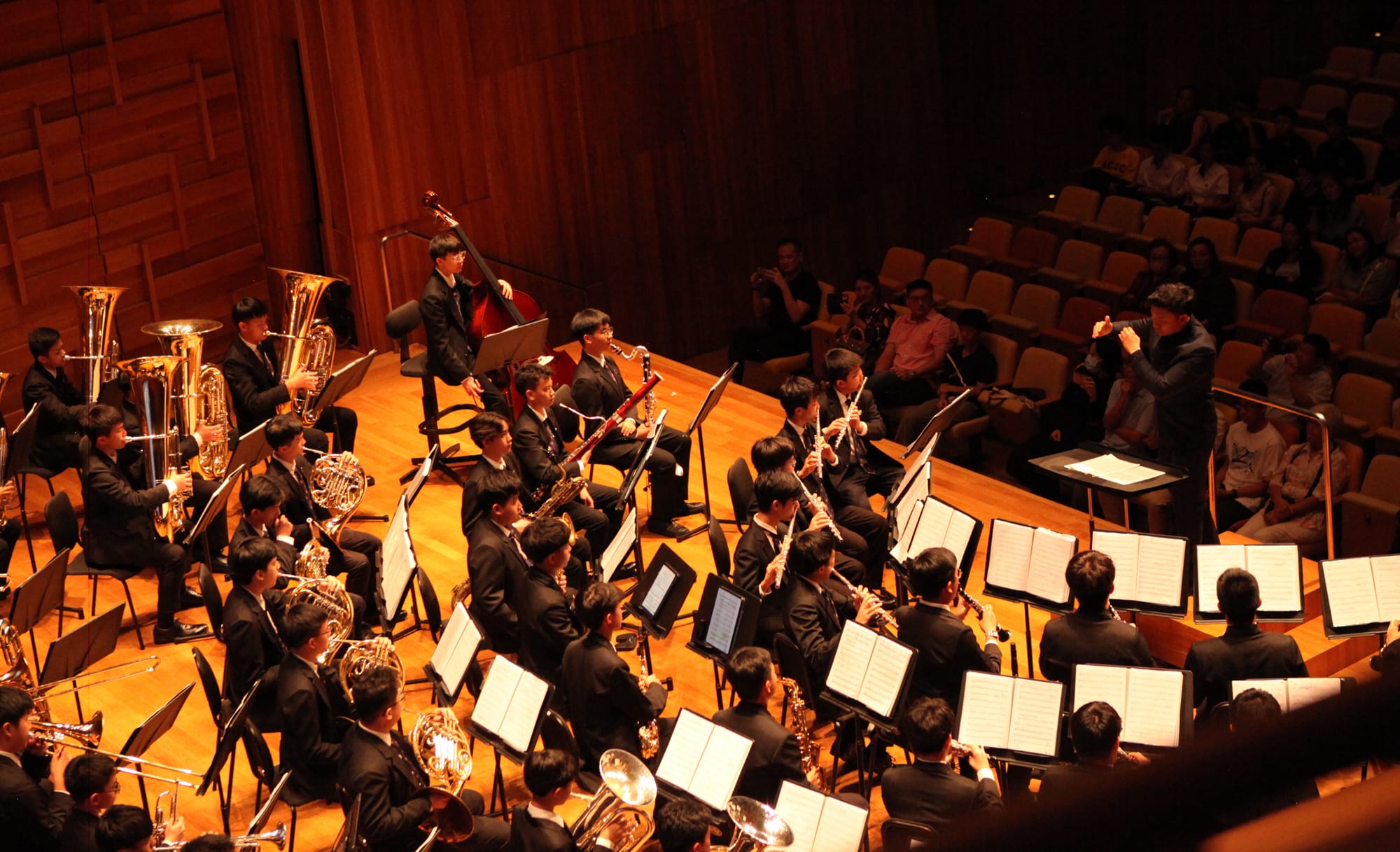 A youth orchestra in black formal attire performs on a wooden concert hall stage, led by a conductor.