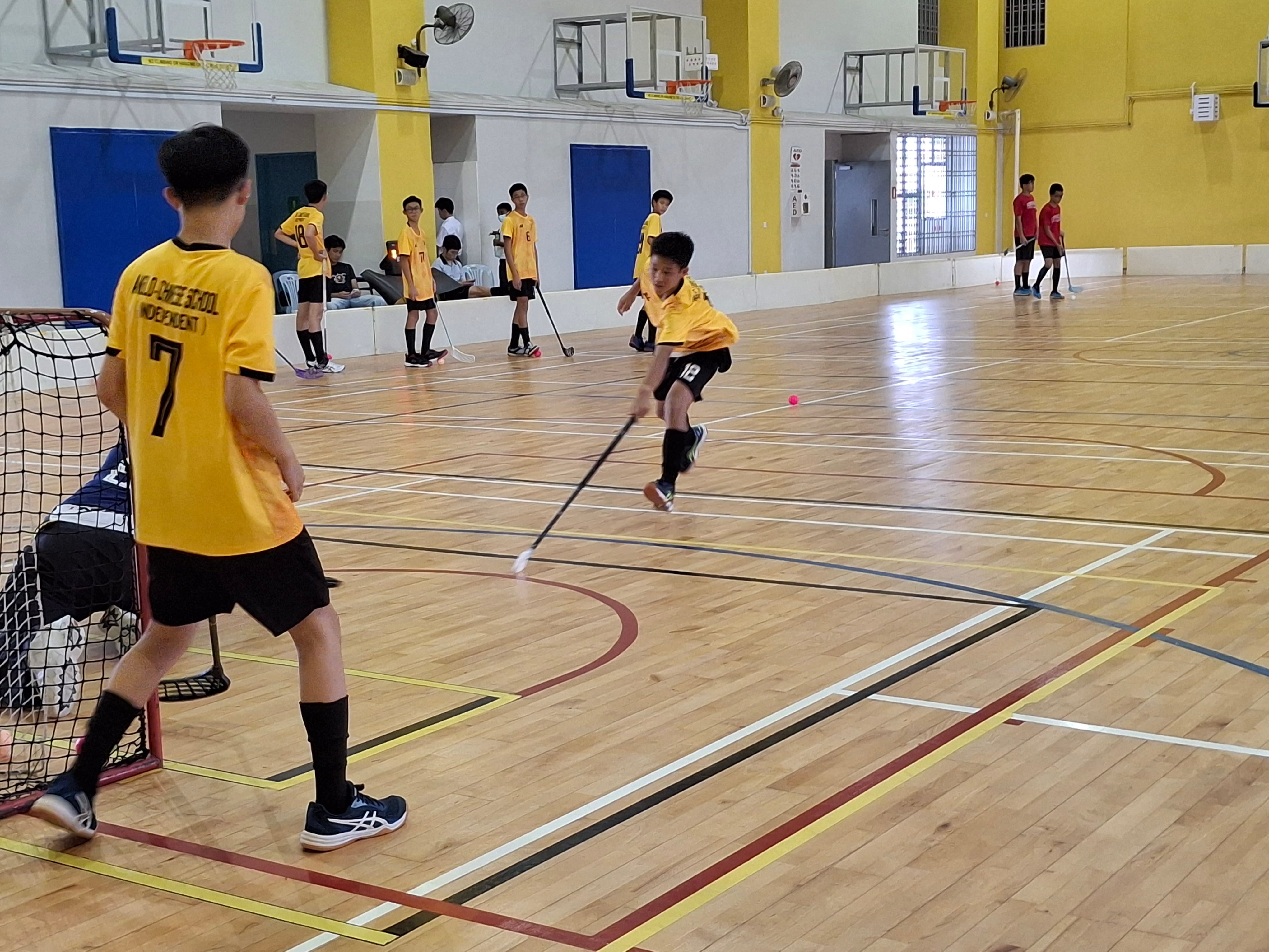 Boys in yellow uniforms playing floorball indoors, one player striking a pink ball toward a goal net.