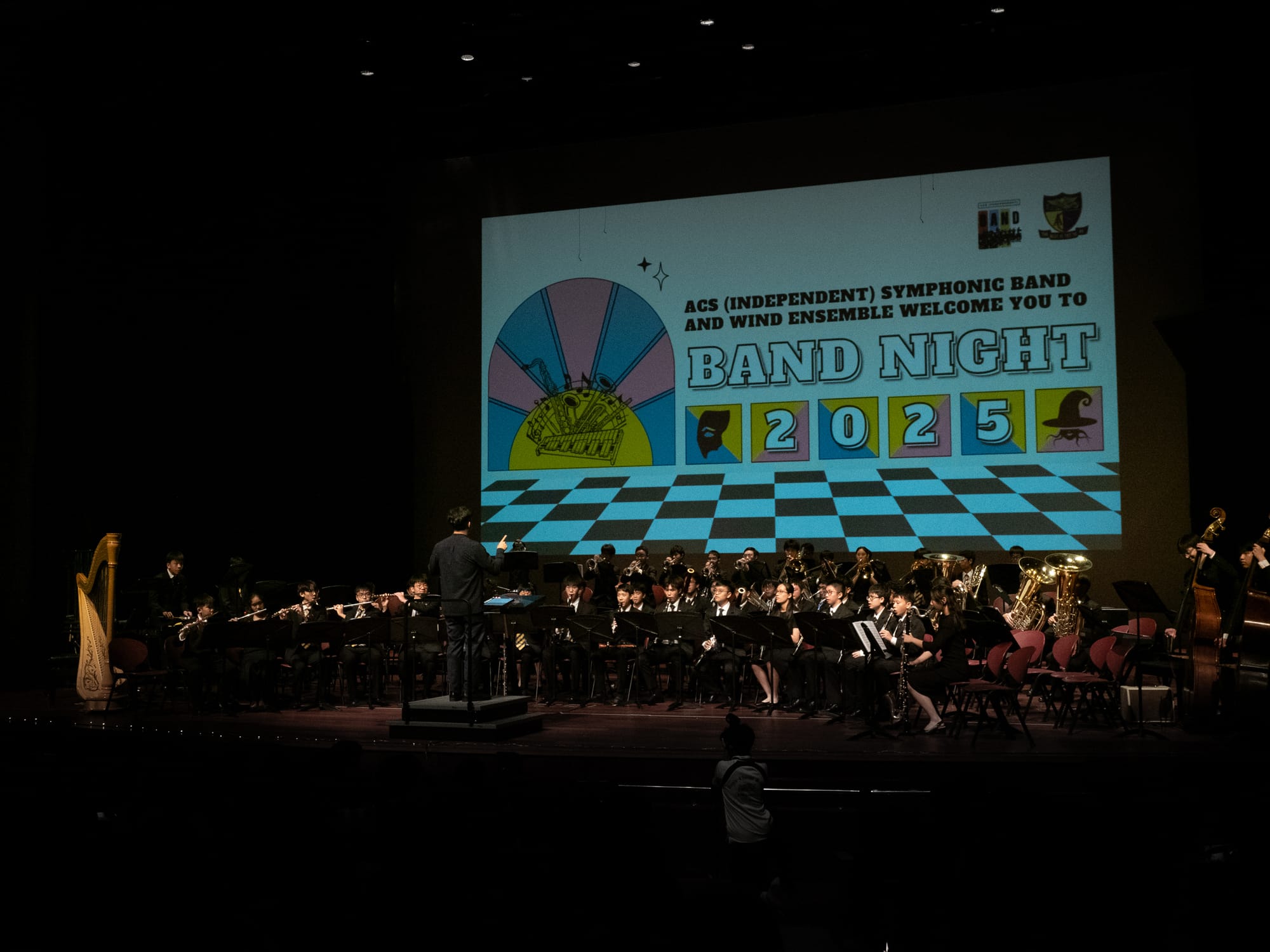 A conductor leads a youth symphonic band on a dark stage at ACS Band Night 2025, with a colorful projected screen behind them.