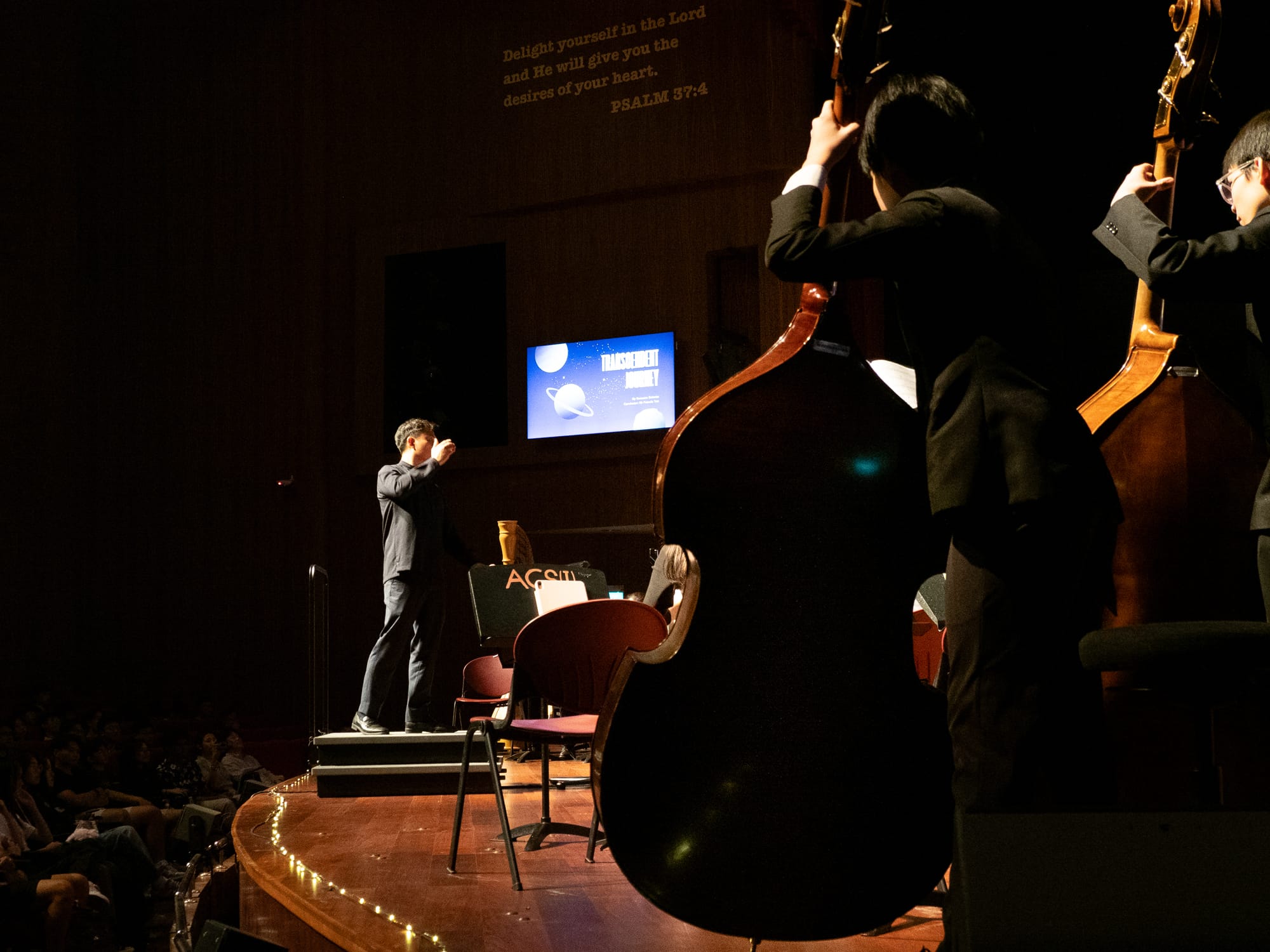 A conductor stands on a dim stage before an orchestra, with double bass players in the foreground and a blue slideshow screen behind.