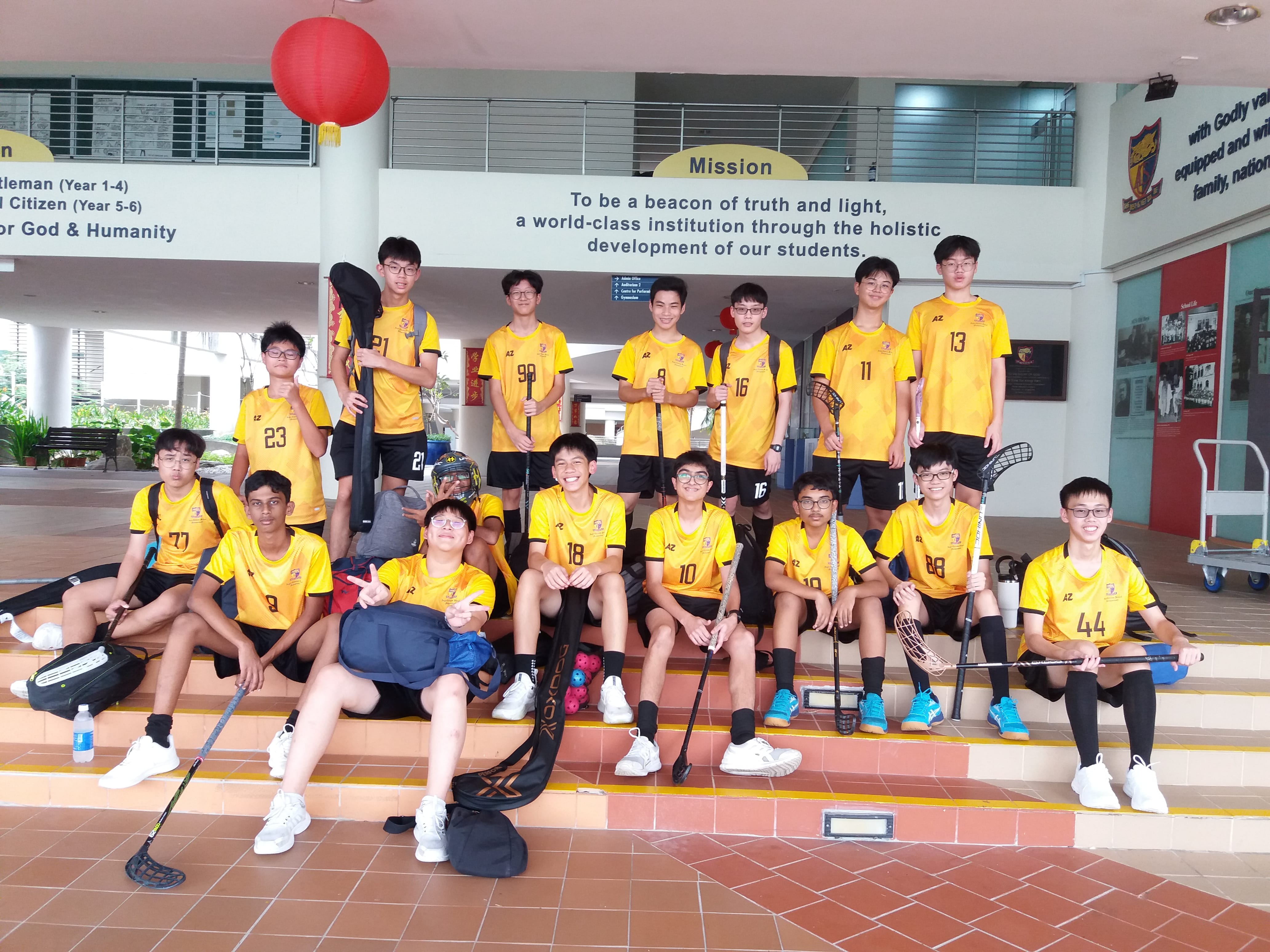 Group of students in yellow jerseys pose on steps with floorball sticks under a mission sign and red lantern