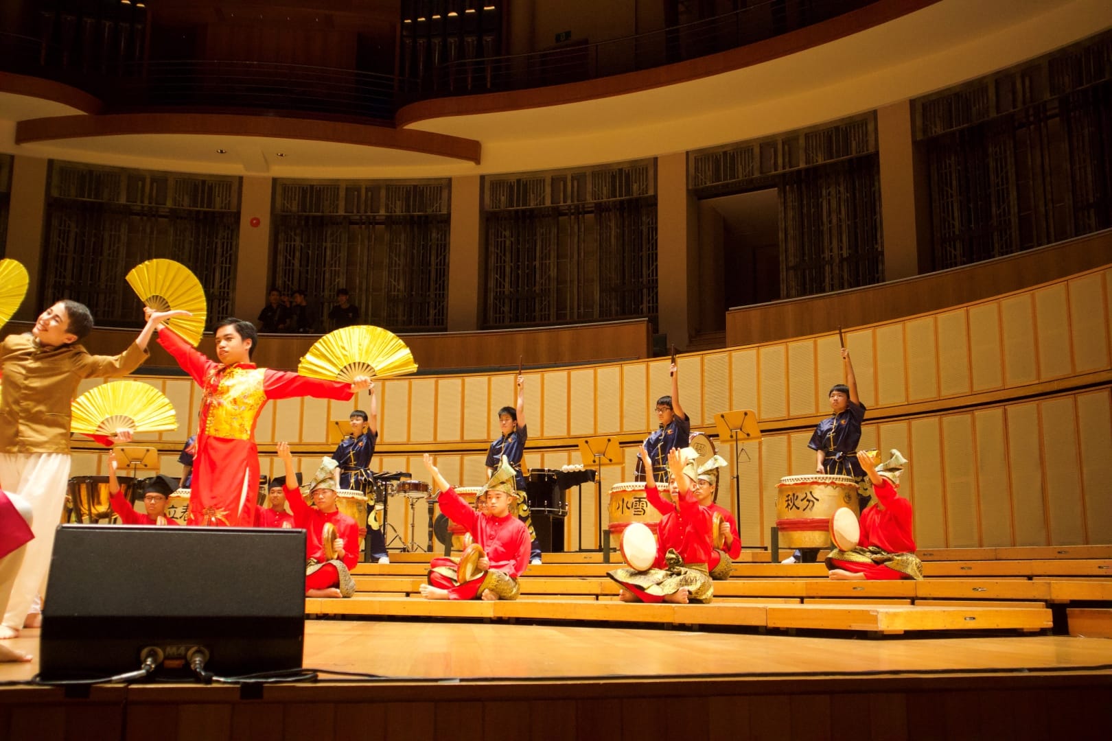 ACS(I) CPAC members perform a combined multicultural showcase on the stage of a professional concert hall, with Chinese fan dancers in red and gold costumes holding large golden fans in the foreground, kompang players in red seated on tiered steps in the middle, and festival drummers in navy uniforms playing large drums on the upper level.