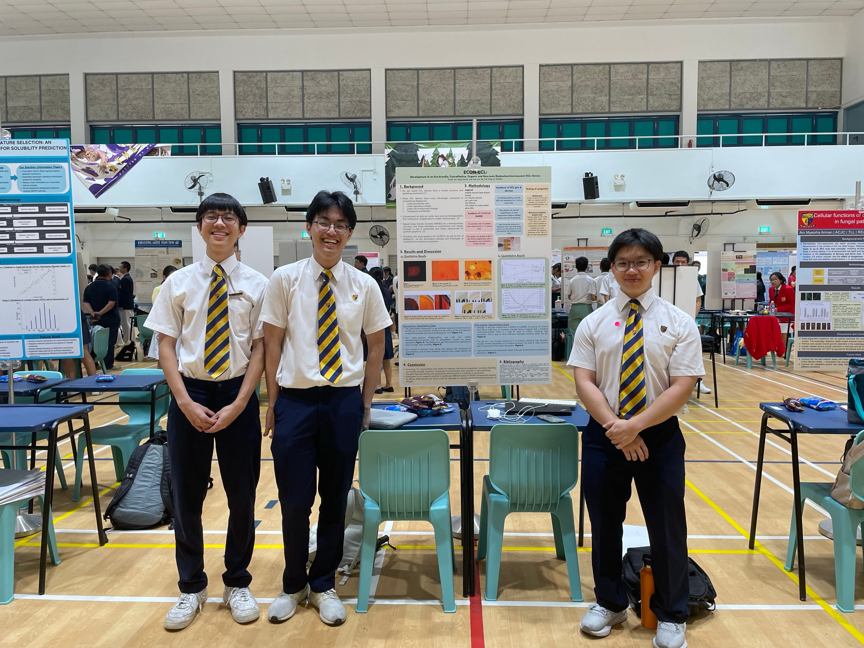Three students in school uniform stand smiling in front of a detailed project poster in a bright, spacious hall. The atmosphere is busy and academic.